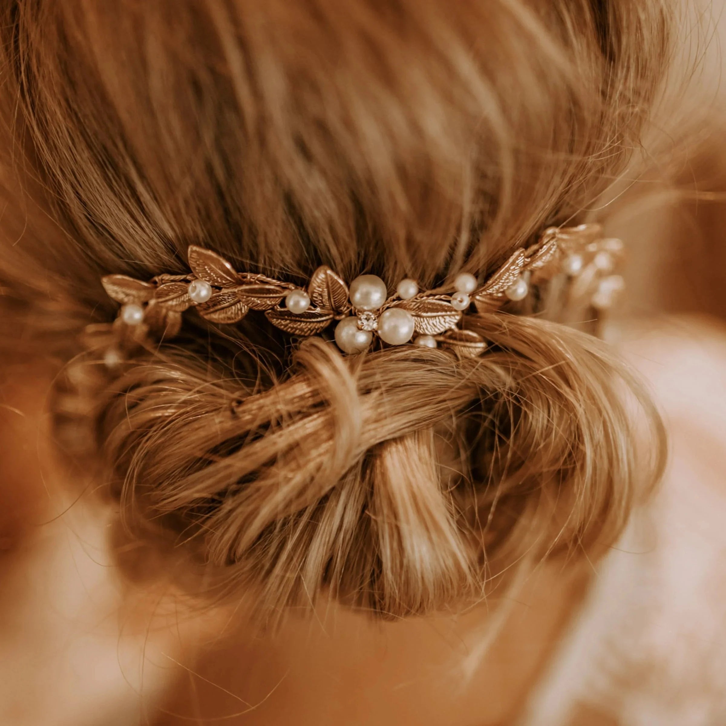 Blondes Haar, gestylt zu einem lockeren, niedrigen Dutt, geschmückt mit einem zarten goldenen Haarteil mit Perlen und kleinen Blattakzenten, was einen weichen, eleganten Look ergibt.