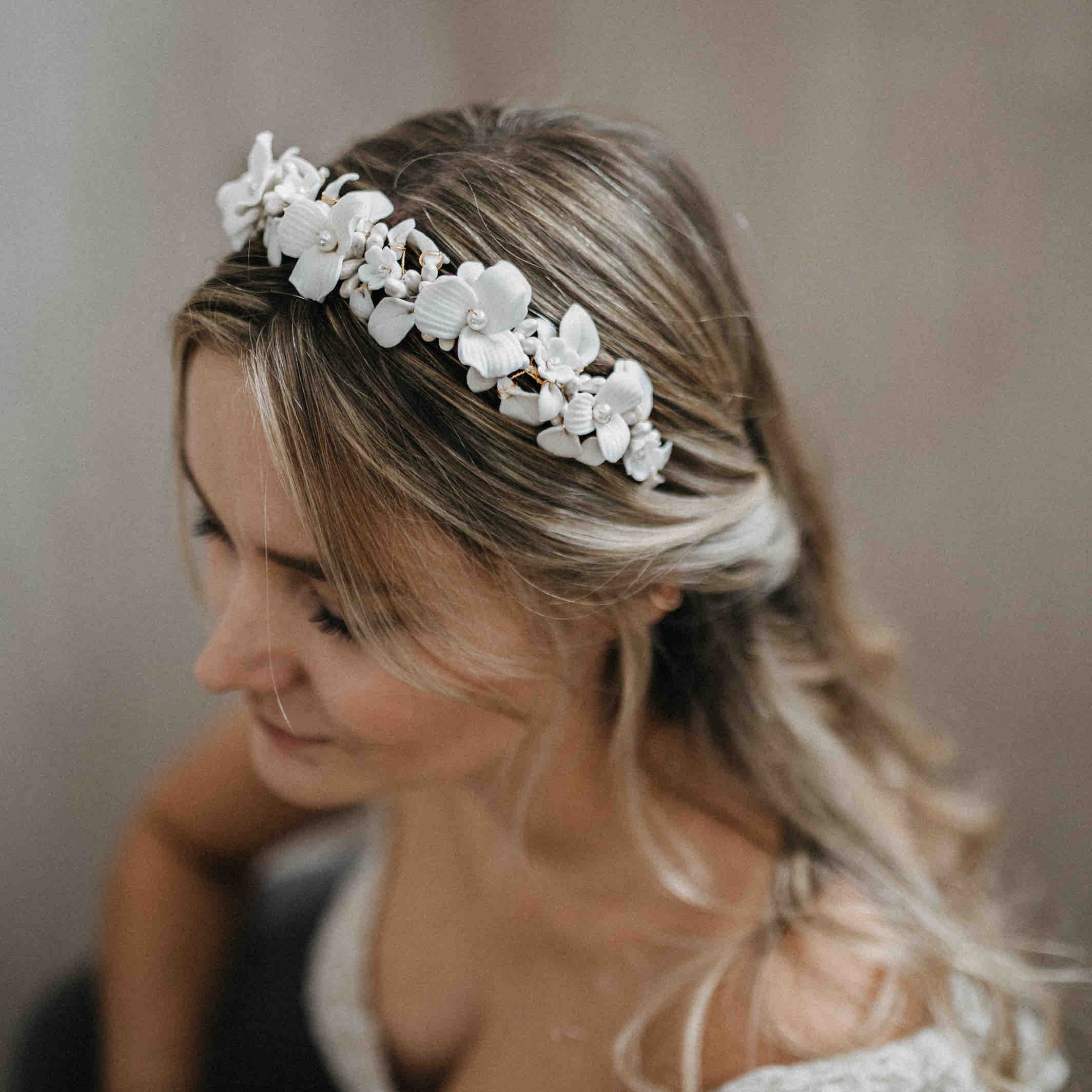Eine Frau mit gewelltem, blondem Haar trägt ein weißes, geblümtes Stirnband. Das Stirnband ist mit zarten, weißen Blumen und kleinen Perlen verziert. Sie blickt nach unten und lächelt sanft. Der Hintergrund ist sanft verschwommen.
