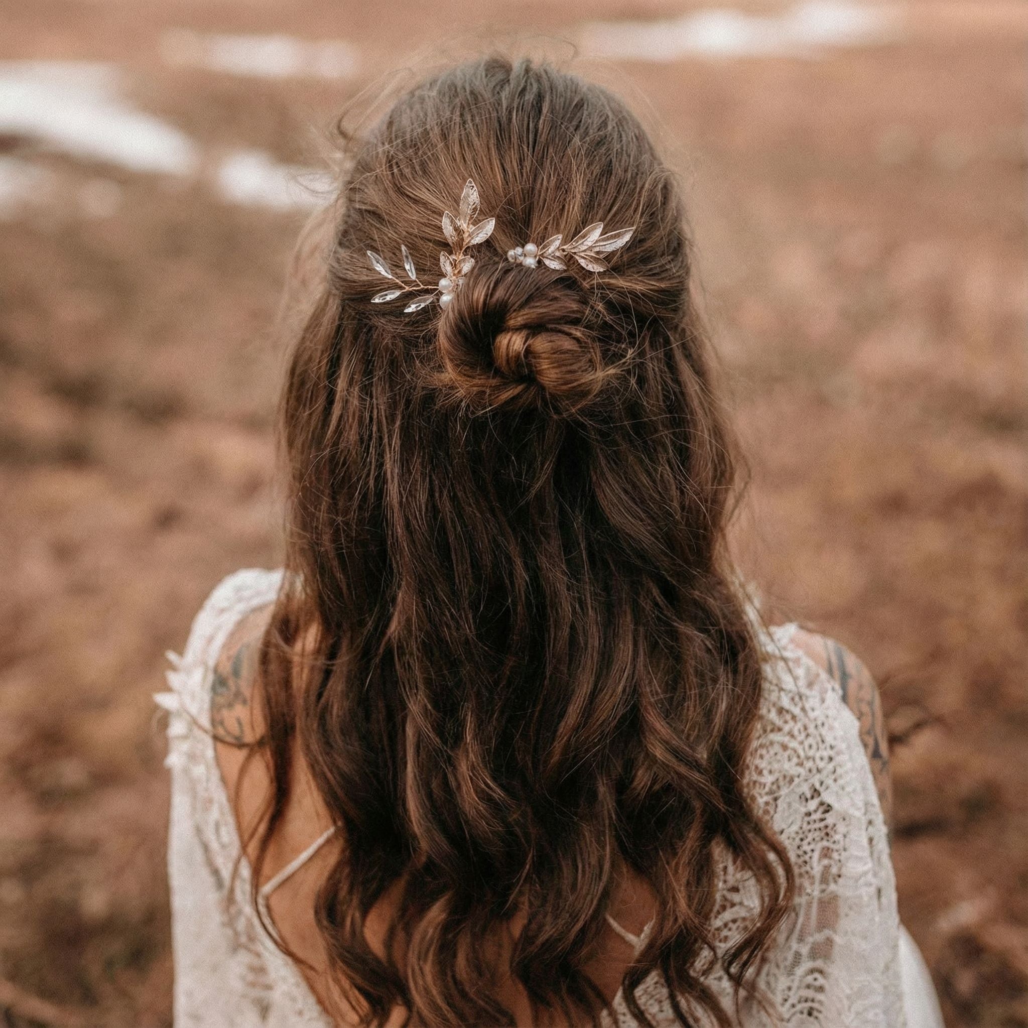 Eine Frau mit langen, gewellten braunen Haaren trägt eine Hochsteckfrisur, die mit einem zarten blattförmigen Haarschmuck verziert ist. Sie steht im Freien, gekleidet in weiße Spitze, vor einem unscharfen, erdigen Hintergrund.