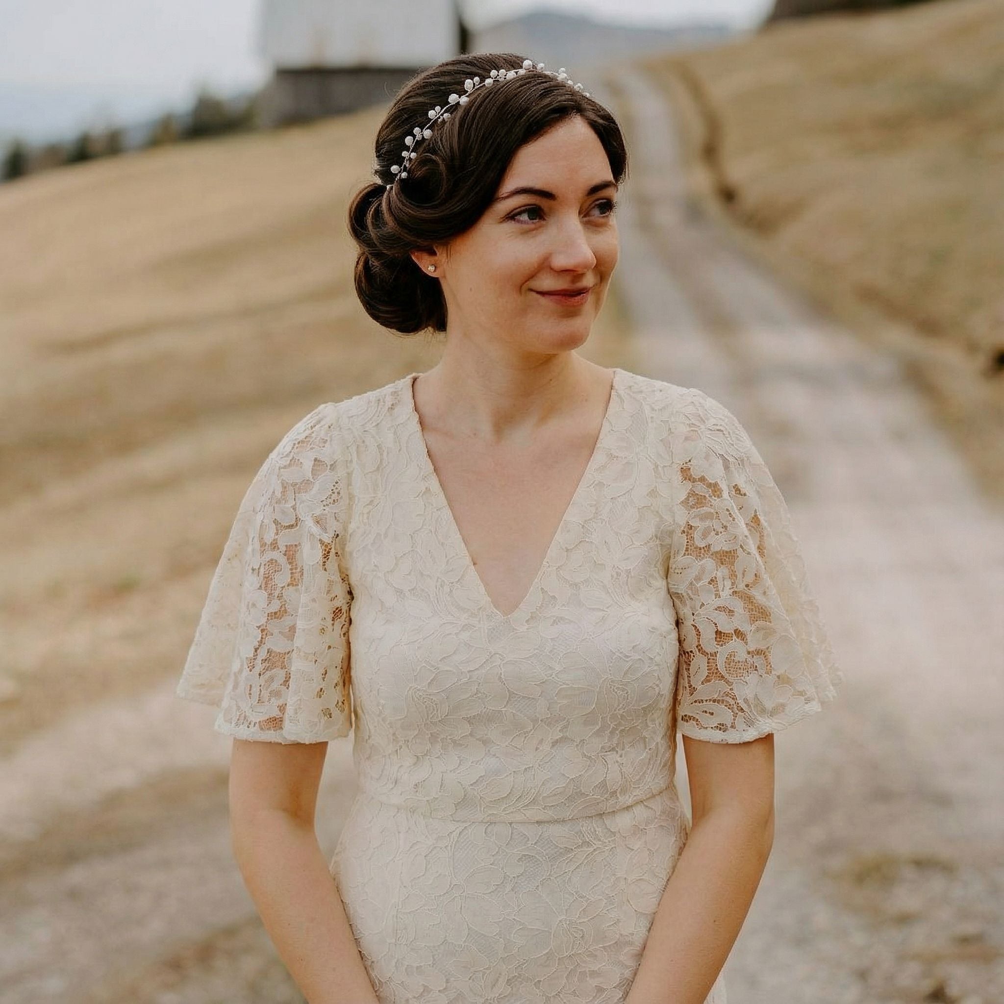 Eine Frau in einem cremefarbenen Spitzenkleid mit Flatterärmeln steht im Freien auf einem ländlichen Feldweg. Sie hat dunkles Haar, das zu einer Hochsteckfrisur mit einem Perlenstirnband frisiert ist, und lächelt sanft und schaut zur Seite. Der Hintergrund ist grasbewachsen mit einer Scheune und bewölktem Himmel.