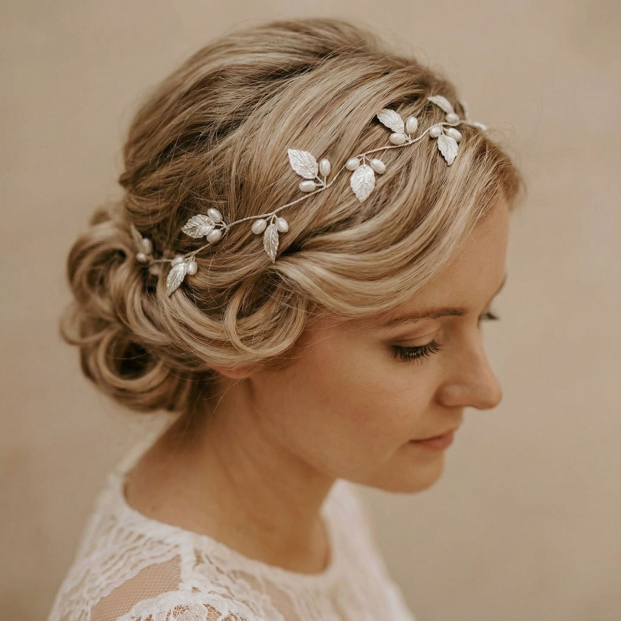 Eine Frau mit blondem Haar, das zu einem niedrigen, eleganten Dutt frisiert ist, trägt ein zartes Stirnband aus Silberblättern. Sie blickt nach links, hat die Augen geschlossen und einen heiteren Ausdruck. Sie trägt ein Spitzenkleid mit komplizierten Mustern. Der Hintergrund hat eine weiche, beige Farbe.