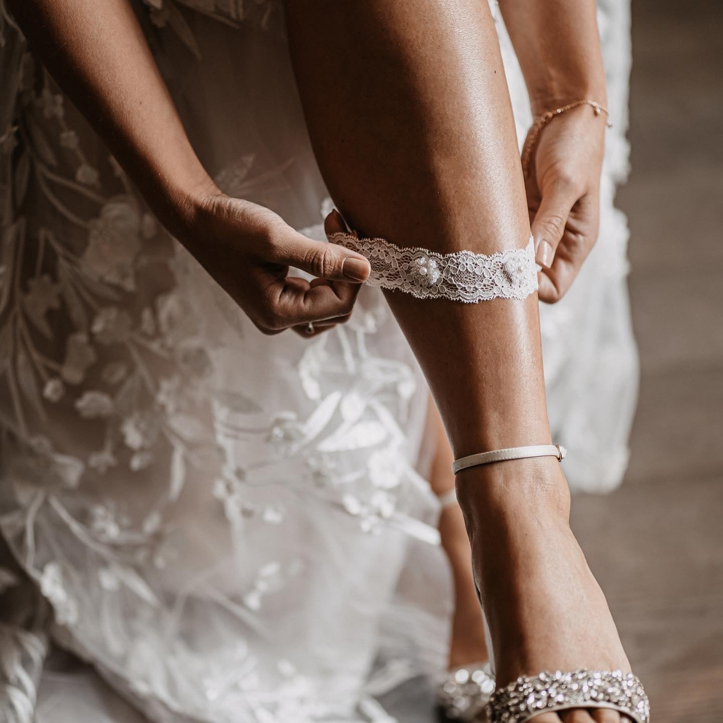 Eine Braut in einem weißen Spitzenkleid und mit Juwelen besetzten Absätzen rückt das Braut Strumpfband Spitze mit Perlen Stickerei von Fräulein Maya auf ihrem Oberschenkel zurecht, wodurch die zarte Spitzen- und Perlenstickerei hervorgehoben wird - ein eleganter Moment für ein Brautaccessoire.