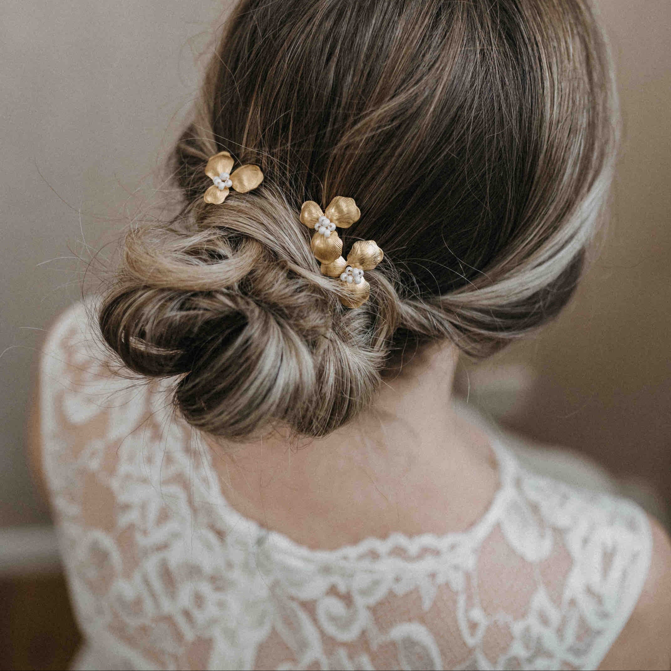 Eine Frau mit braunem Haar in einem niedrigen, strukturierten Dutt trägt Haarnadeln mit goldenen Blüten (3 Stück) von Fräulein Maya. Von hinten in einem weißen Spitzenkleid betrachtet, kommen die elegante Frisur und die filigrane Spitze schön zur Geltung.