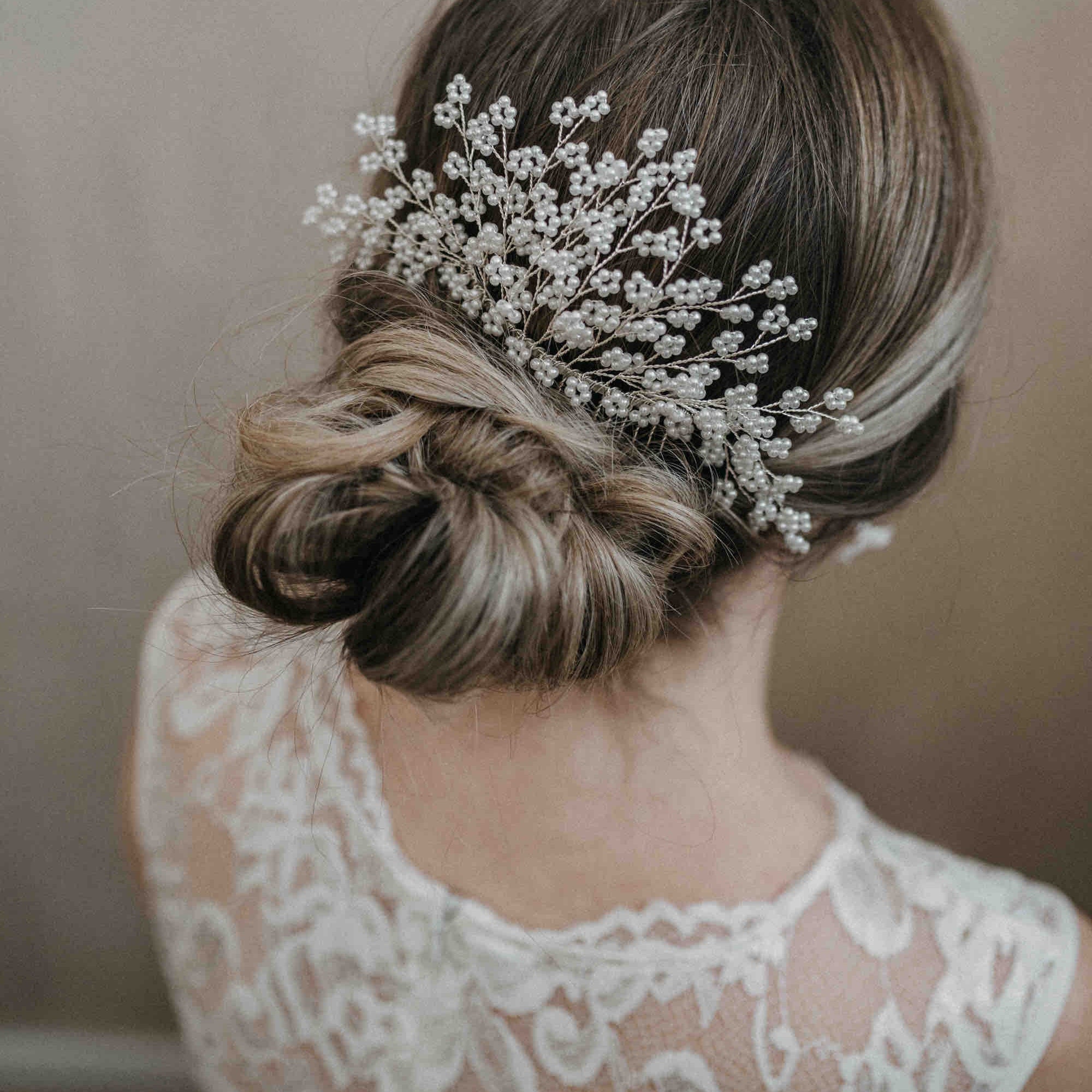 Eine Frau mit brünettem Haar, das zu einer niedrigen, lockeren Hochsteckfrisur gestylt ist, die mit dem Haarkamm Braut mit Perlen (groß) von Fräulein Maya verziert ist. Sie trägt ein weißes, mit Blumen gemustertes Spitzenkleid, von hinten gesehen vor einem neutralen Hintergrund.