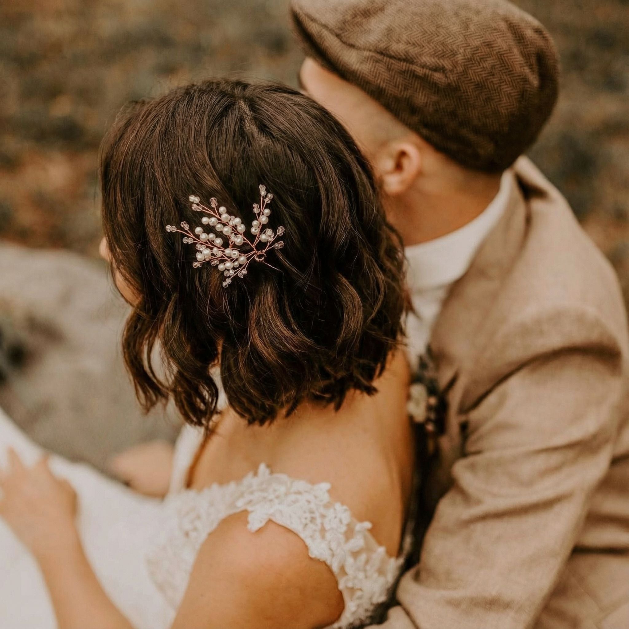 Ein Paar sitzt eng beieinander im Freien. Die Frau in einem weißen schulterfreien Spitzenkleid und mit Perlenschmuck im Haar wendet sich ab. Der Mann, der einen hellbraunen Anzug und eine braune, flache Kappe trägt, umarmt sie von hinten. Der Hintergrund ist unscharf und erdig.