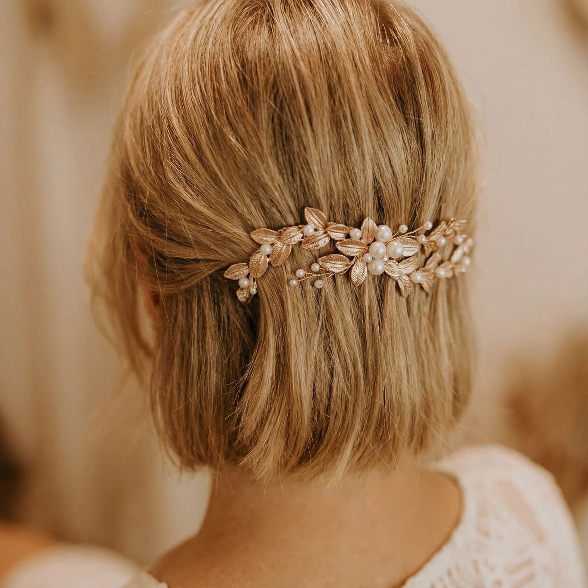 Eine Frau mit kurzem, gewelltem blondem Haar, das zu einer lockeren Hochsteckfrisur frisiert ist. Ihr Haar ist mit einem dekorativen goldenen Haarteil mit Perlen- und Blattakzenten geschmückt. Sie trägt ein cremefarbenes Spitzenoberteil, und der Hintergrund ist sanft verschwommen.