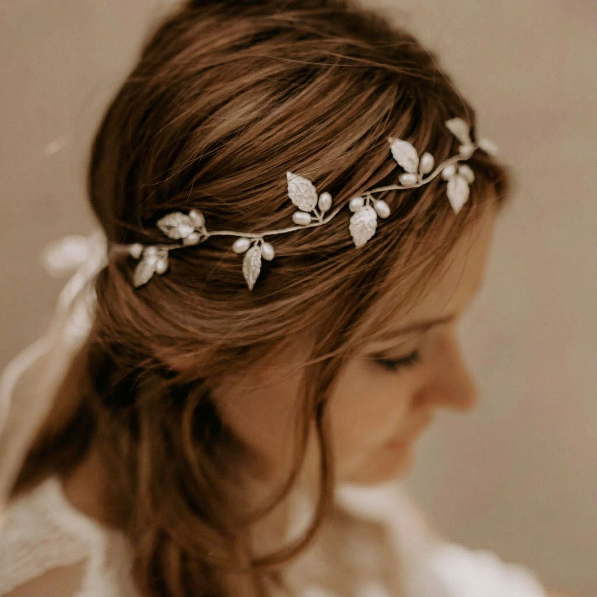 Eine Frau mit hellbraunem Haar trägt ein zartes Stirnband aus Blattsilber und Perlen. Ihr Haar ist in lockeren Wellen gestylt, und sie blickt nach unten, teilweise unscharf. Der Hintergrund ist sanft verschwommen und schafft eine sanfte, romantische Atmosphäre.