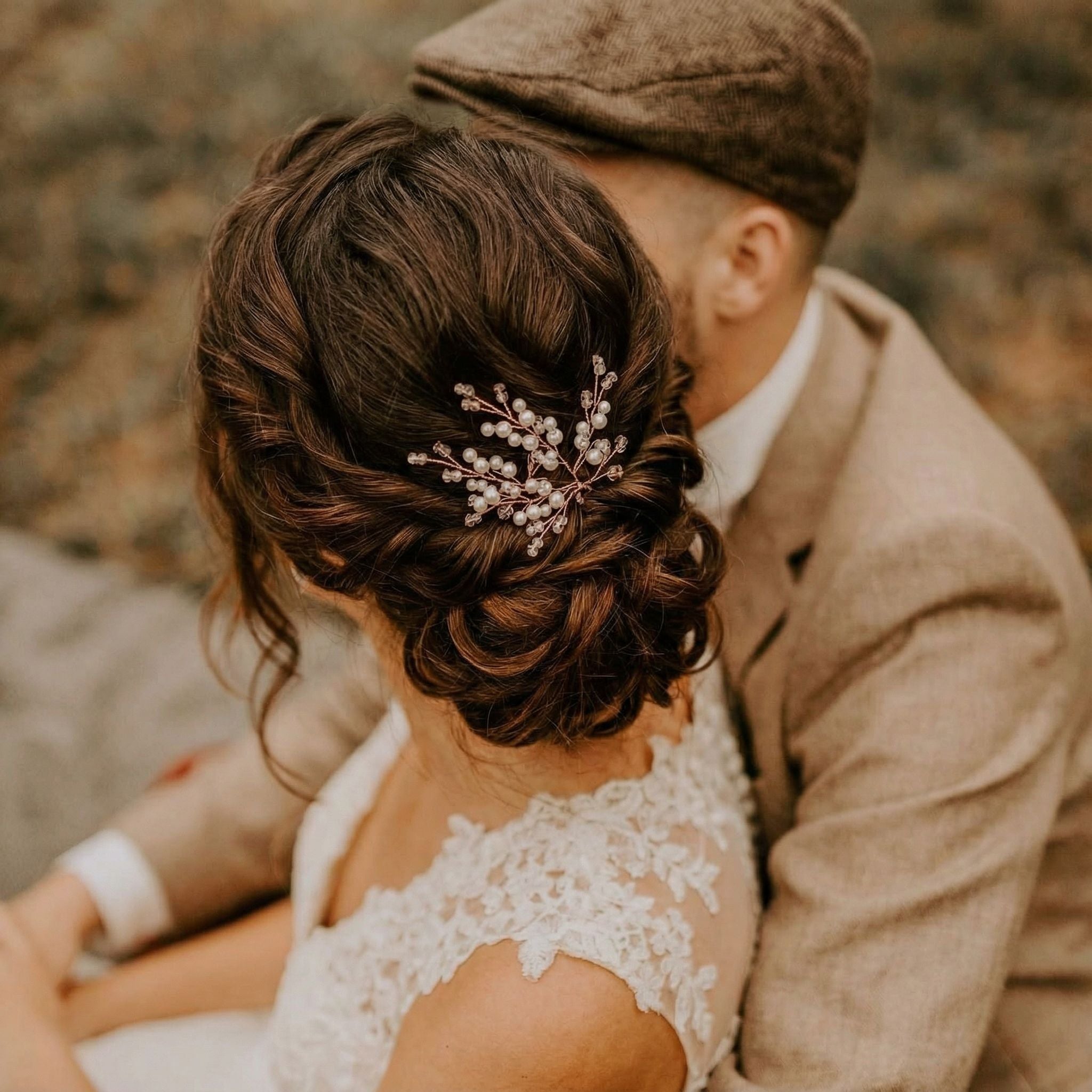 Eine Frau mit einer lockeren Hochsteckfrisur, die mit einem Haarteil aus Perlen und Kristallen geschmückt ist, sitzt eng mit einem Mann in einem beigen Anzug und einer flachen Kappe zusammen. Sie trägt ein Hochzeitskleid aus Spitze und ihre Gesichter sind teilweise verdeckt, während sie einen intimen Moment im Freien teilen.