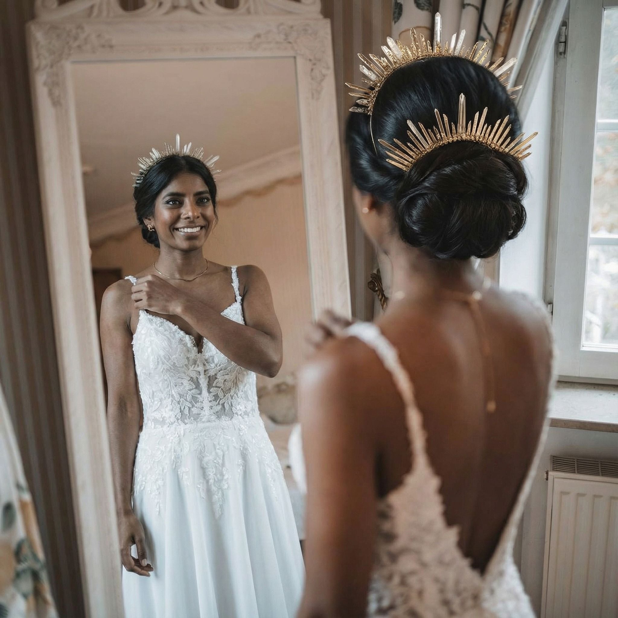 Eine Frau in einem weißen Spitzenhochzeitskleid und einem goldenen Stachelkopfschmuck steht lächelnd vor einem großen, verzierten Spiegel und betrachtet ihr Spiegelbild. Ihr Haar ist zu einem eleganten Dutt frisiert, und durch ein nahes Fenster fällt sanftes Licht ein.