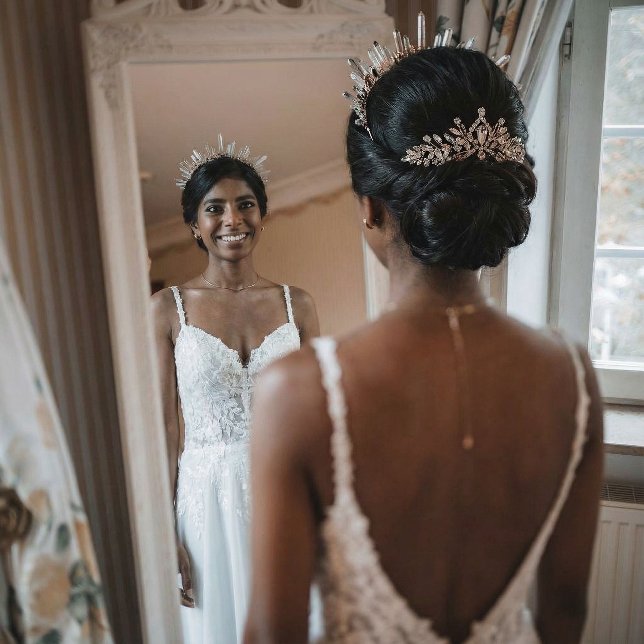 Eine Braut mit dunklem Haar in einer kunstvollen Hochsteckfrisur, geschmückt mit einer juwelenbesetzten Tiara, lächelt ihr Spiegelbild in einem großen Spiegel an. Sie trägt ein weißes Spitzenhochzeitskleid mit dünnen Trägern und steht in einem sanft beleuchteten, eleganten Raum mit cremefarbenen Wänden und Vorhängen.