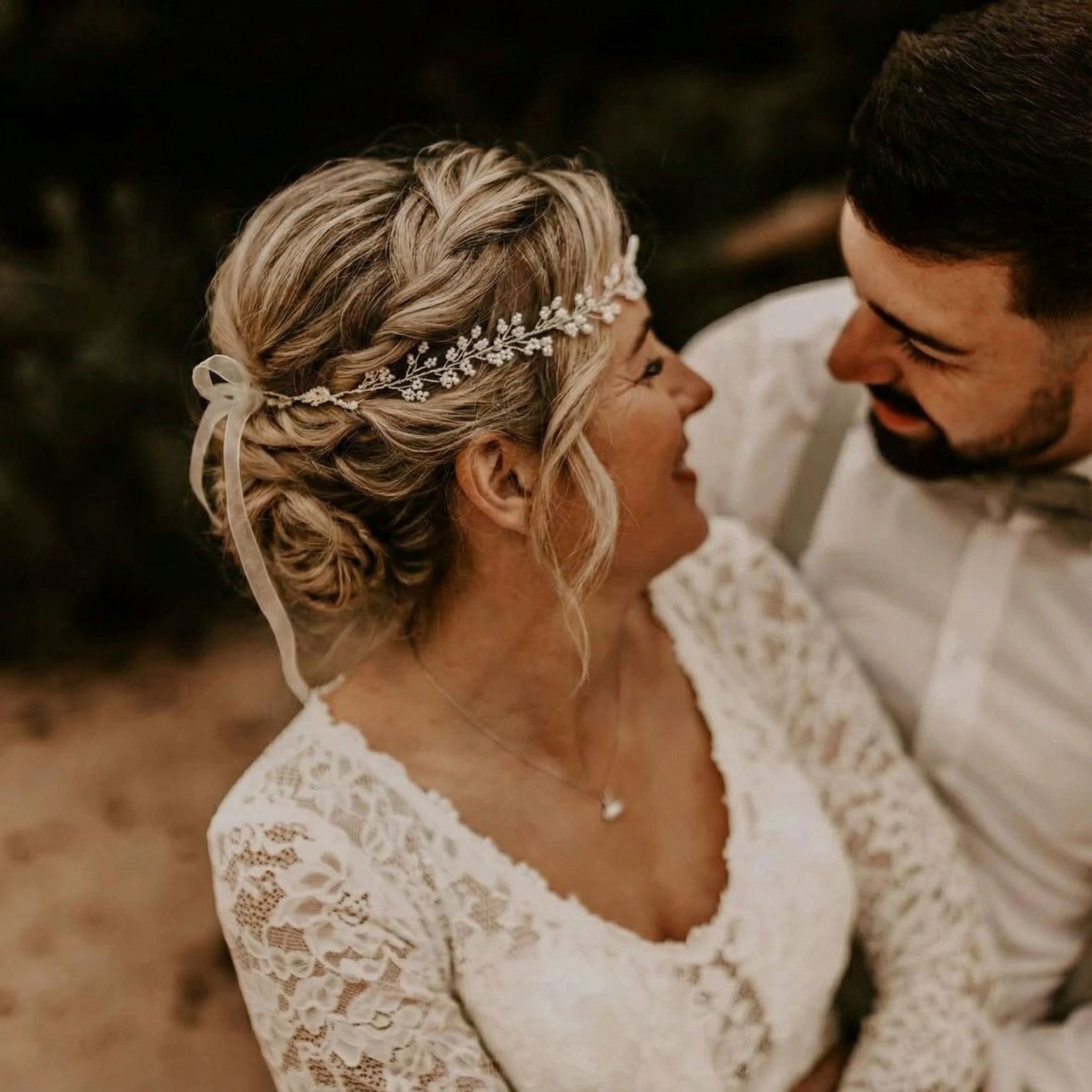 Eine Braut mit geflochtenen blonden Haaren, die mit einem zarten Blumenkopfschmuck und einem Spitzenkleid geschmückt sind, lächelt einen bärtigen Bräutigam mit weißem Hemd und Fliege herzlich an. Sie stehen dicht beieinander und teilen einen intimen, freudigen Moment im Freien.
