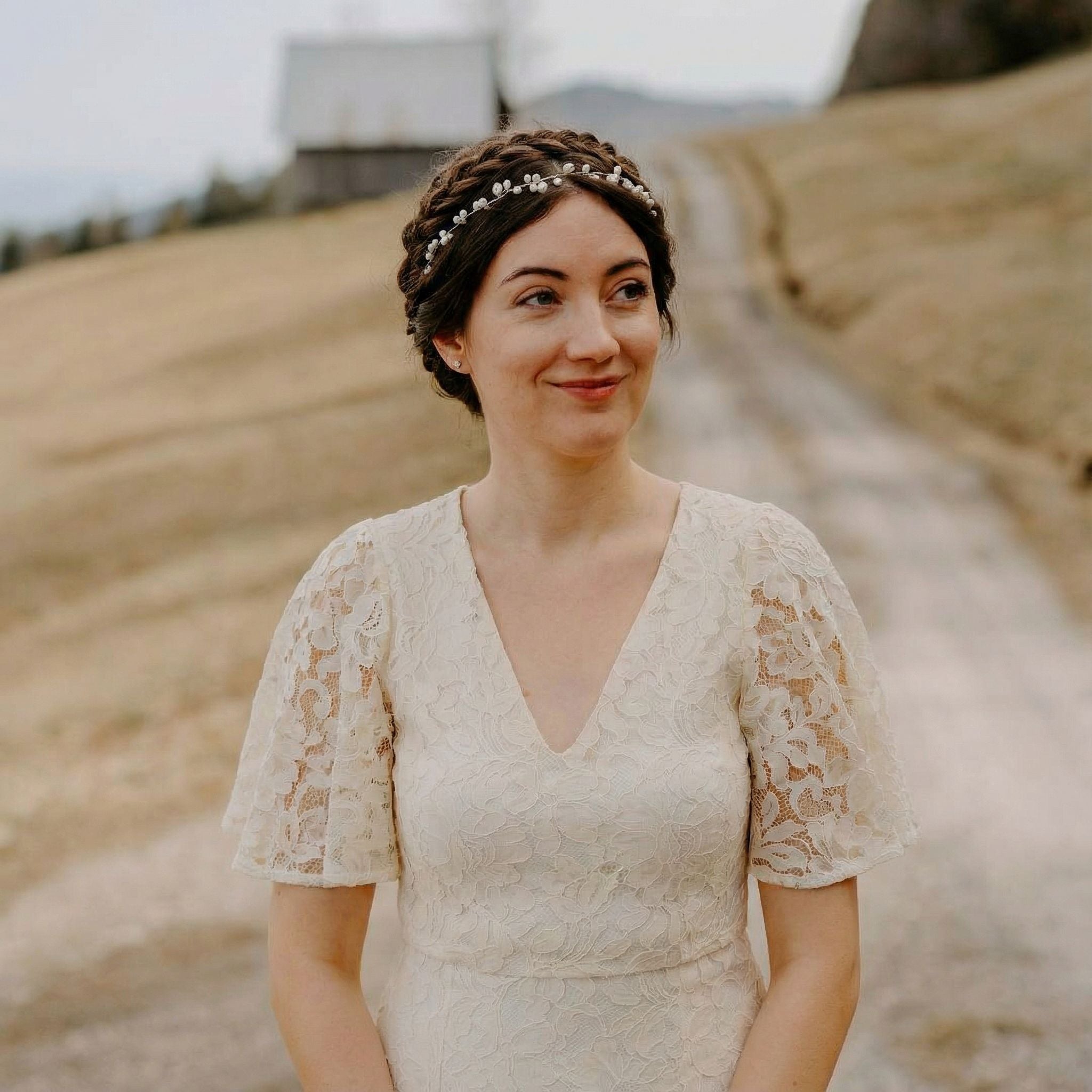 Eine Frau in einem Spitzenhochzeitskleid mit Flatterärmeln und einem Perlenstirnband steht auf einem Feldweg. Sie hat dunkles, geflochtenes Haar, lächelt sanft und blickt leicht zur Seite. Der Hintergrund zeigt ein unscharfes Feld und ein rustikales Gebäude.