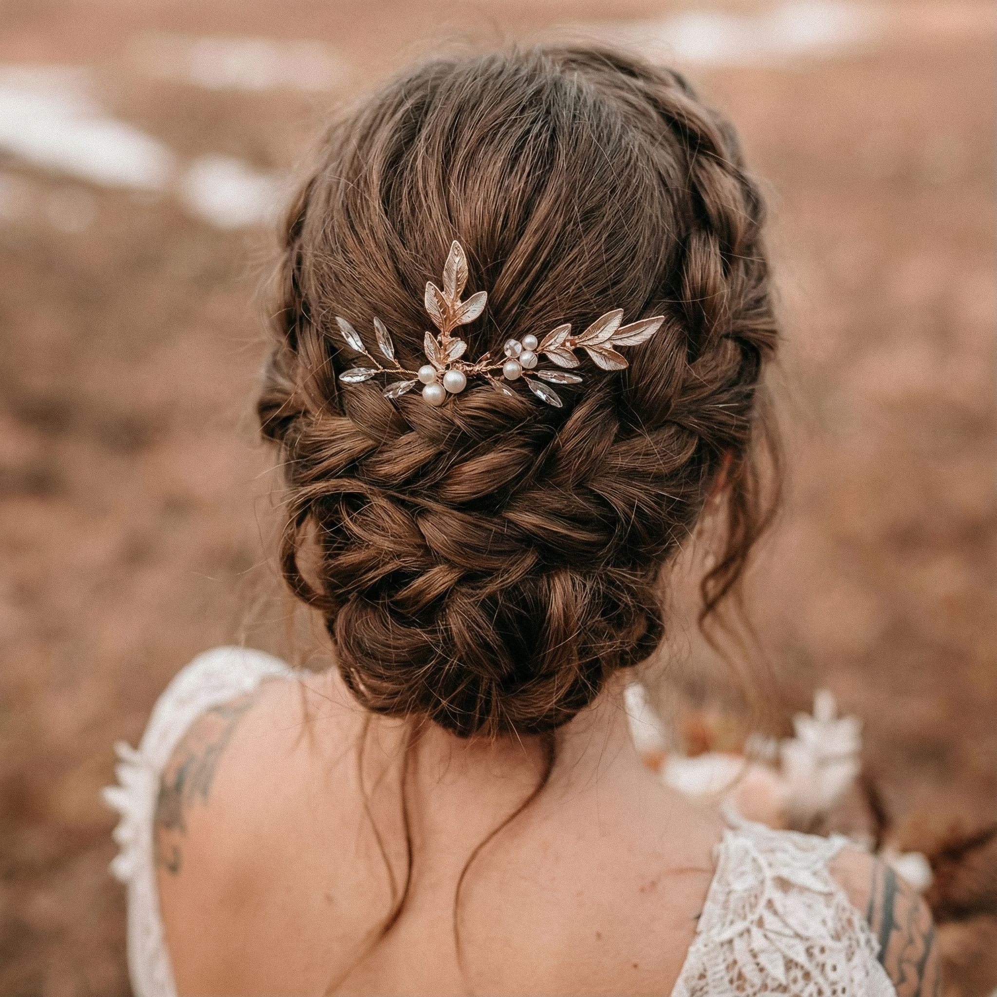 Eine Frau mit braunem Haar, das zu einer eleganten geflochtenen Hochsteckfrisur frisiert und mit einem zarten goldenen Haarschmuck mit Perlen und Blättern verziert ist. Sie trägt ein spitzenbesetztes Kleid im Vintage-Stil. Der Hintergrund ist im Freien und leicht unscharf.