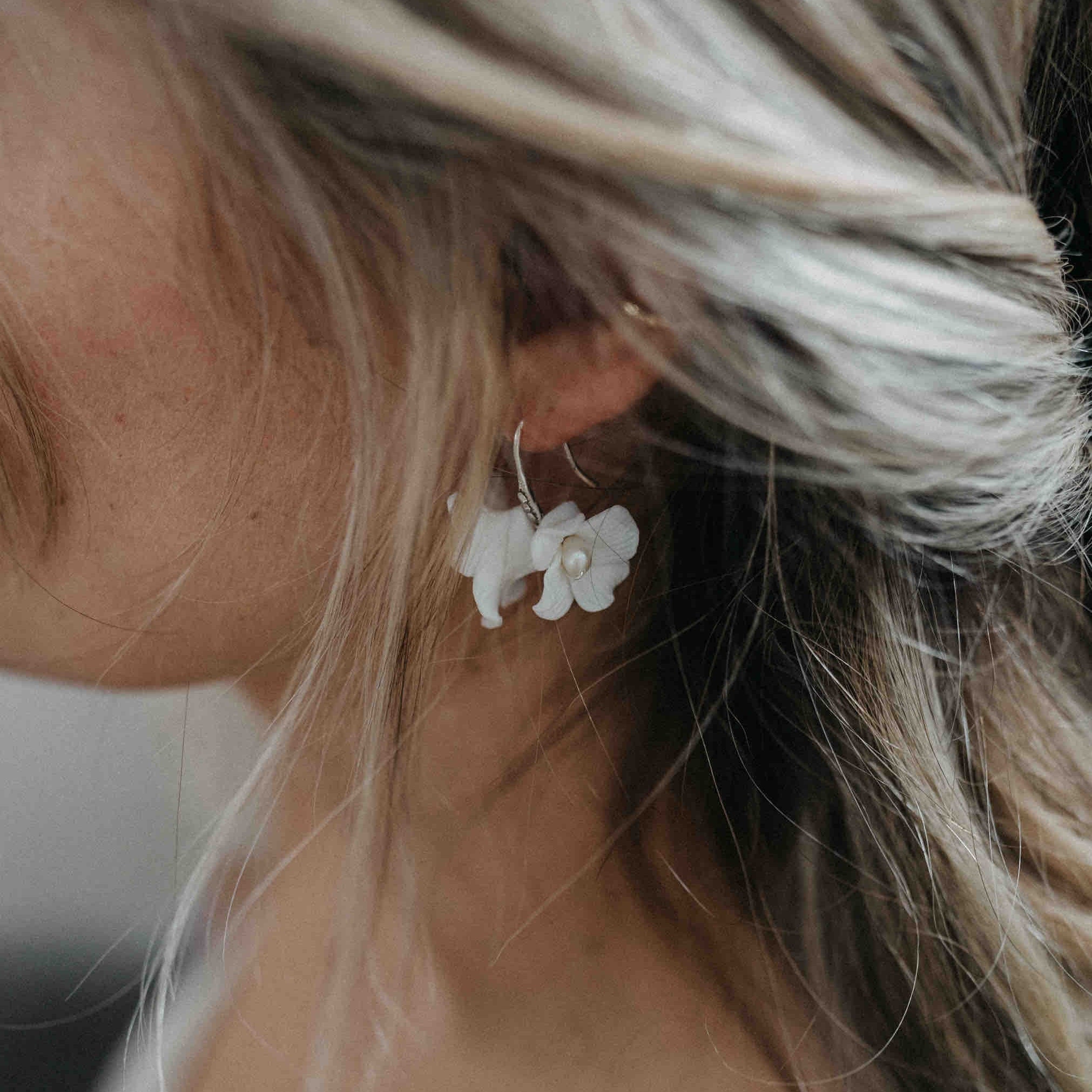 Nahaufnahme des Ohrs einer Person mit gewelltem blondem Haar im Boho-Braut-Stil, die Fräulein Maya Braut Ohrringe mit Keramikblüten und einen kleinen Reif darüber trägt, während das Licht ihre Haut und die weichen, natürlichen Haartöne hervorhebt.