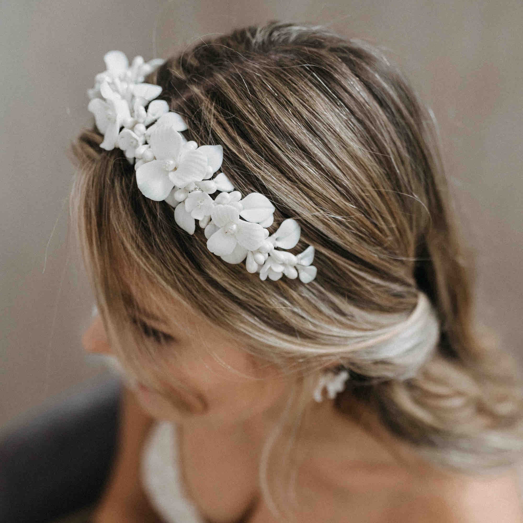 Eine Frau mit lockeren blonden Wellen trägt ein schulterfreies Spitzenkleid. Ihre Hochzeitsfrisur ist mit der Krone Braut mit Keramik Blüten von Fräulein Maya und passenden Ohrringen geschmückt, die von oben gezeigt werden, um ihre Frisur in Szene zu setzen.