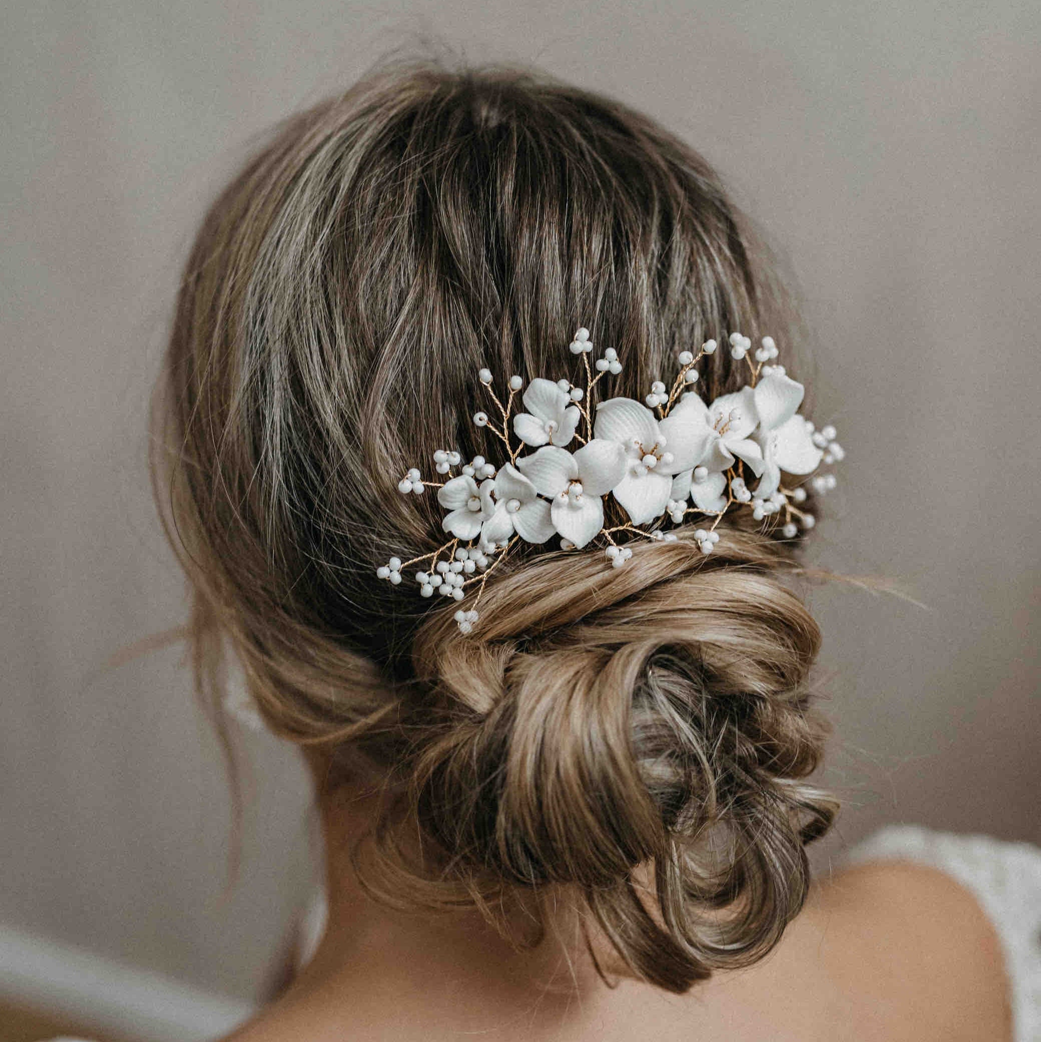 Eine Frau mit hellbraunem Haar in einem strukturierten Dutt trägt den Fräulein Maya Keramik Haarkamm mit Blumen und Perlen, der ihrem Brautlook Eleganz und Romantik verleiht. Der sanft verschwommene Hintergrund unterstreicht die Szene.