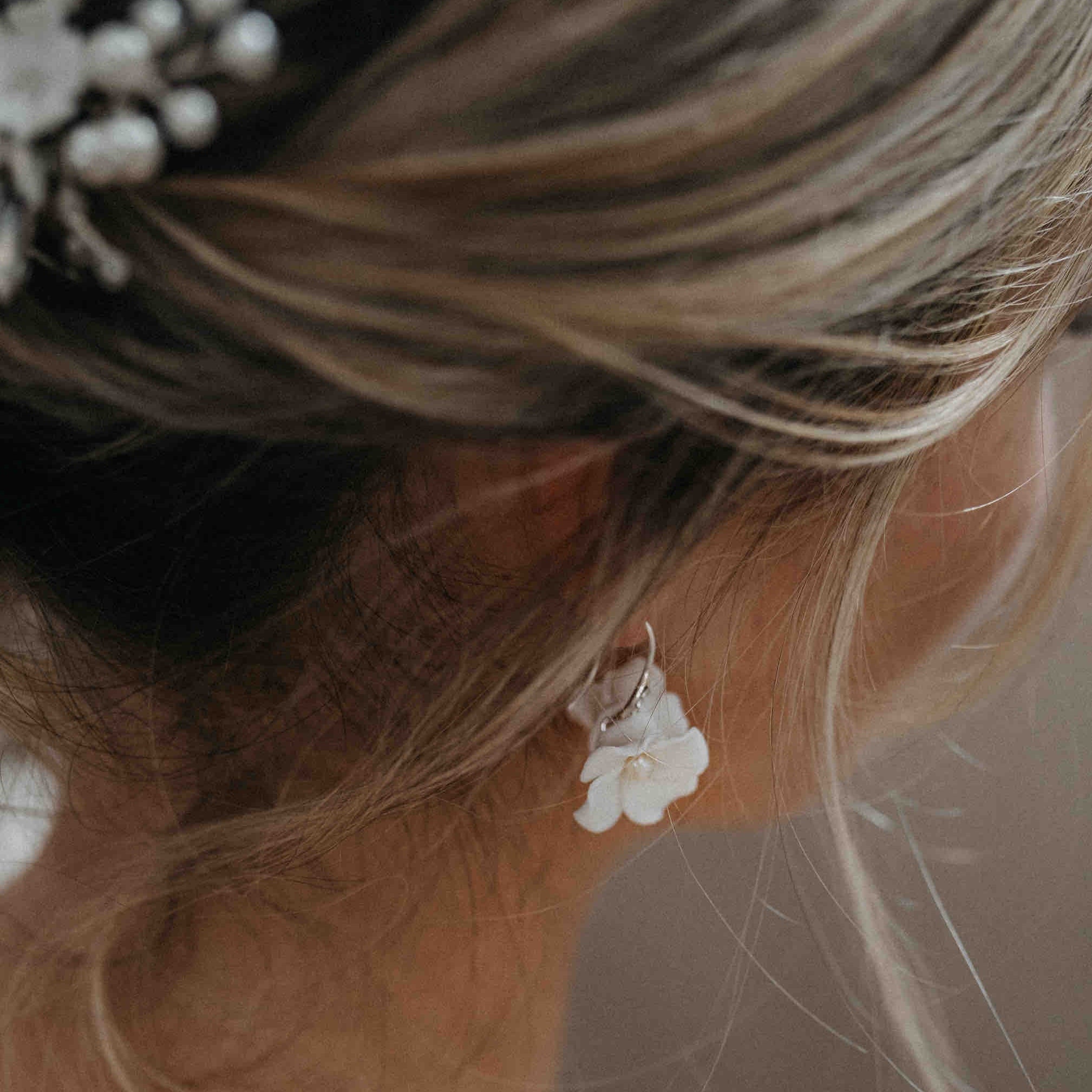 Nahaufnahme der blonden Brautfrisur einer Frau mit Perlenhaarnadeln, die die "Braut Ohrringe mit Keramik Blüten" von Fräulein Maya trägt. Weiches Licht unterstreicht den romantischen Boho-Braut-Look vor einem leicht verschwommenen Hintergrund.
