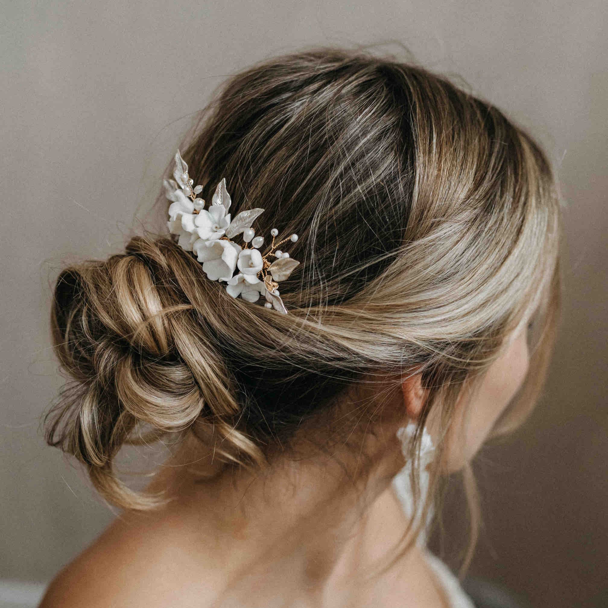 Eine Frau mit blondem Haar, das zu einem lockeren, niedrigen Dutt gestylt ist, trägt den Fräulein Maya Haarkamm mit Keramik Blumen und Blätter, der mit zarten Keramikblumen und goldenen Blättern verziert ist. Sie wendet sich ab und präsentiert ihre elegante Frisur vor einem sanft verschwommenen Hintergrund.