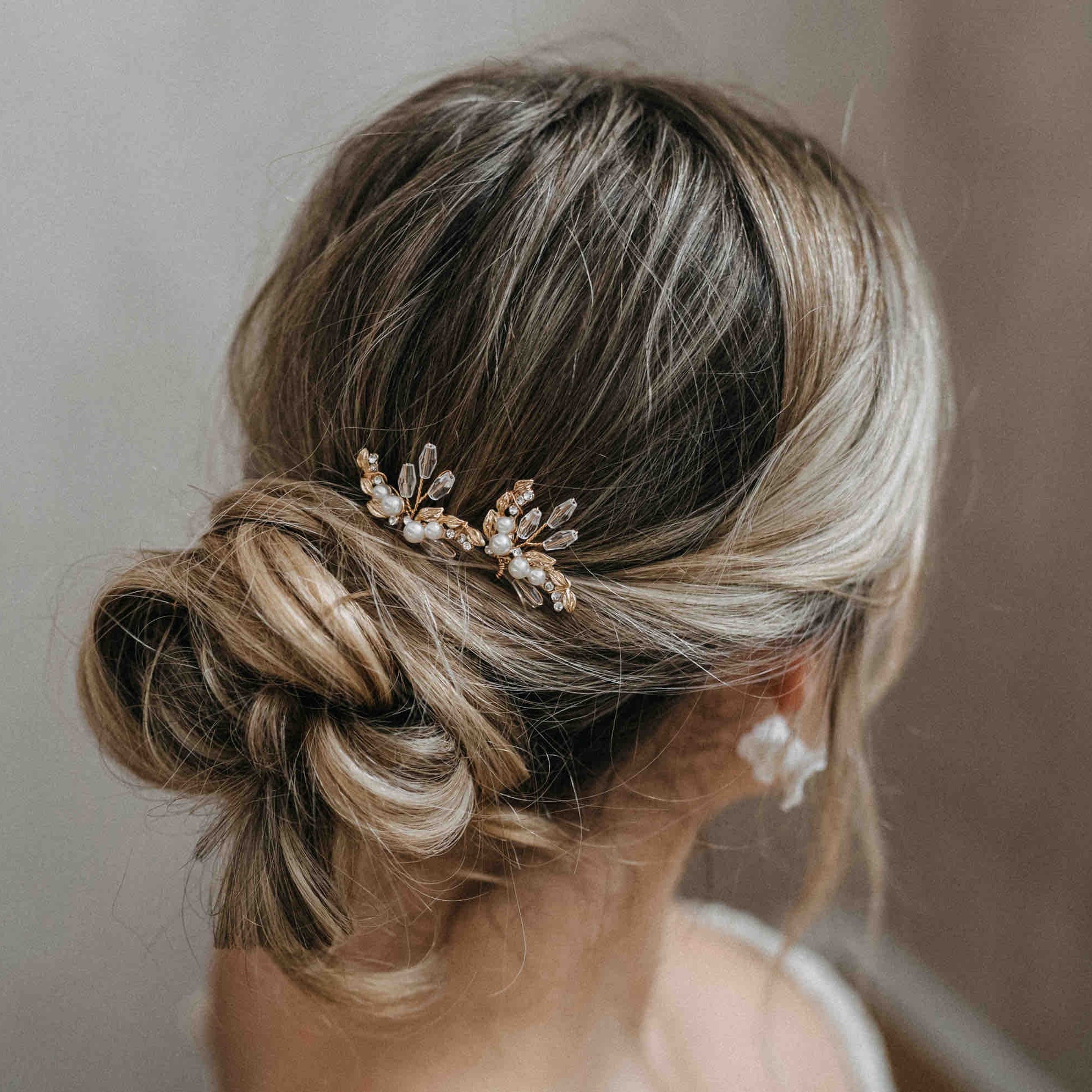 Eine Frau mit hellbraunem Haar in einem eleganten tiefen Dutt trägt Fräulein Maya Kristall Haarnadeln mit funkelnden Steinen (3 Stück), die ihr Glanz verleihen. Sie trägt einen weißen Blumenohrring und ein weißes Oberteil vor einem sanft verschwommenen Hintergrund.