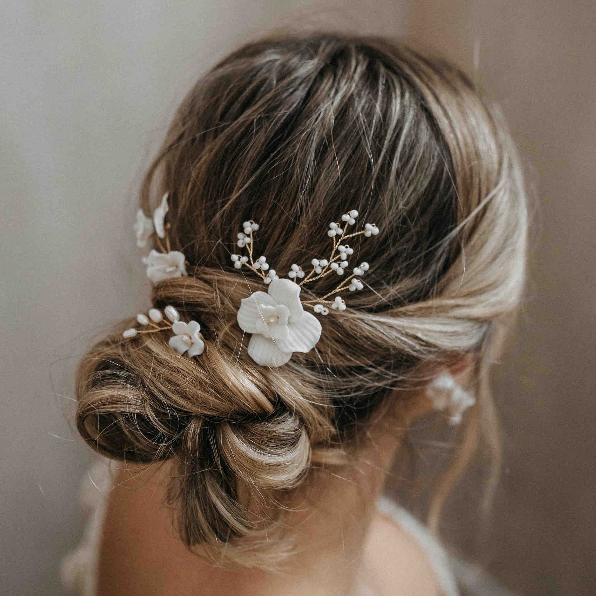 Eine Frau mit blondem Haar in einem lockeren tiefen Dutt trägt Fräulein Maya Haarnadeln mit Keramik Blüten und Perlen (3 Stück), die ihre Brautfrisur mit romantischen weißen Blumen und Perlen ergänzen. Ein unscharfer Hintergrund unterstreicht die elegante Frisur.