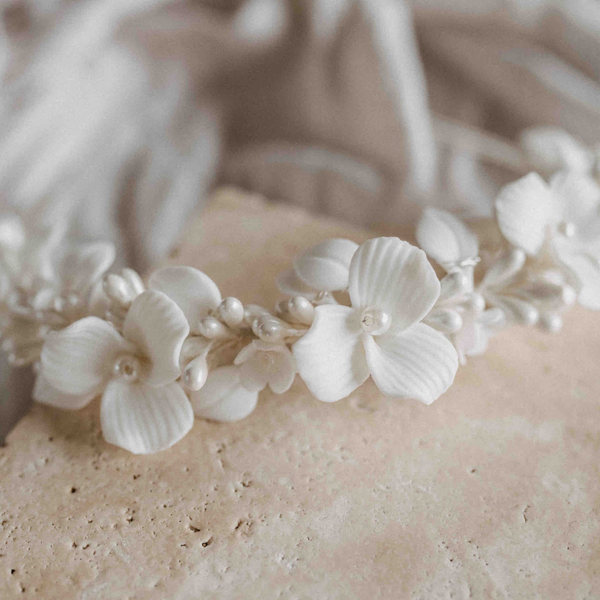 Die Krone Braut mit Keramik Blüten von Fräulein Maya ist ein zartes Stirnband, perfekt für die Brautfrisur, verziert mit weißen handgefertigten Keramikblumen und perlenartigen Perlen. Es wird auf hellbeigem Marmor mit einem weichen, neutral getönten Stoffhintergrund präsentiert.