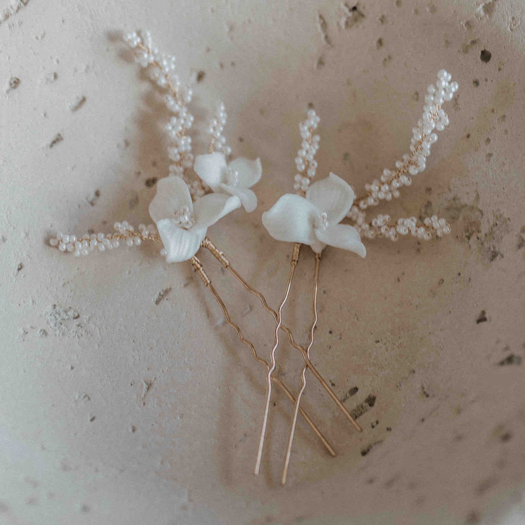 Zwei Keramik-Harnadeln mit Blumen und feinen Perlen von Fräulein Maya (2 Stück) liegen auf einer strukturierten beigen Oberfläche. Die goldenen Stiele und weißen Blüten verleihen dem Arrangement eine weiche, elegante Ausstrahlung.