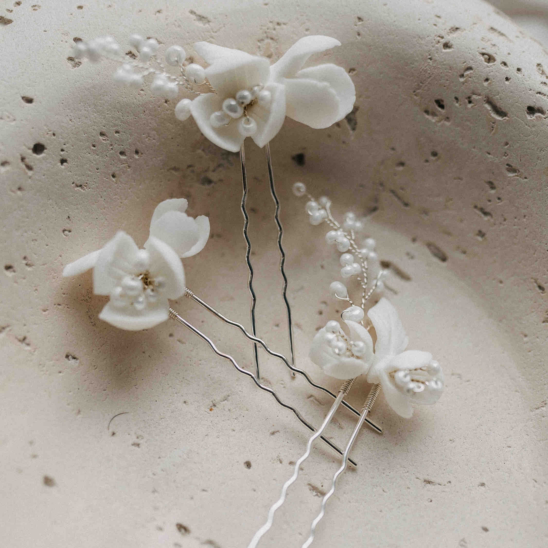 Drei Haarnadeln mit großen Keramik Blumen von Fräulein Maya (3 Stück) liegen auf einer strukturierten, cremefarbenen Steinfläche und präsentieren die eleganten botanischen Haaraccessoires stilvoll.