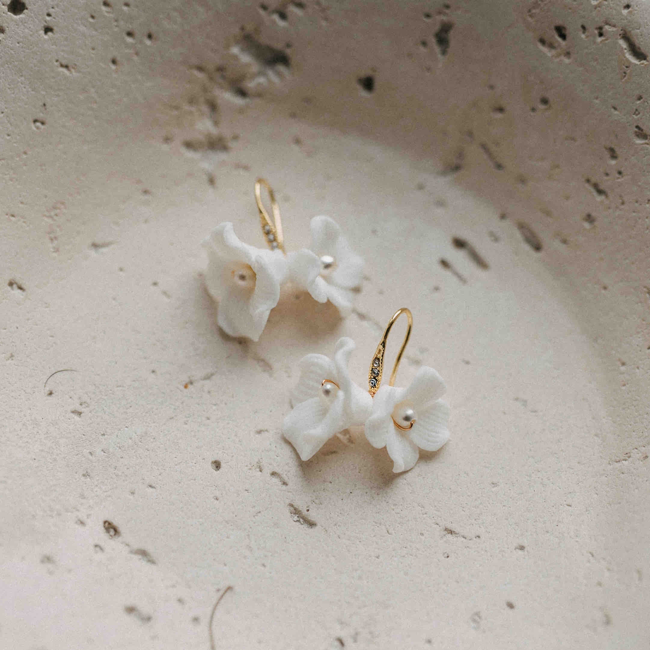 Die Braut Ohrringe mit Keramik Blüten von Fräulein Maya präsentieren weiße Blumen mit goldenen Creolen und Perlenzentrum auf beigem Keramik. Sanftes Licht betont ihr organisches Design - ideal für Boho-Bräute, die natürliche Eleganz lieben.