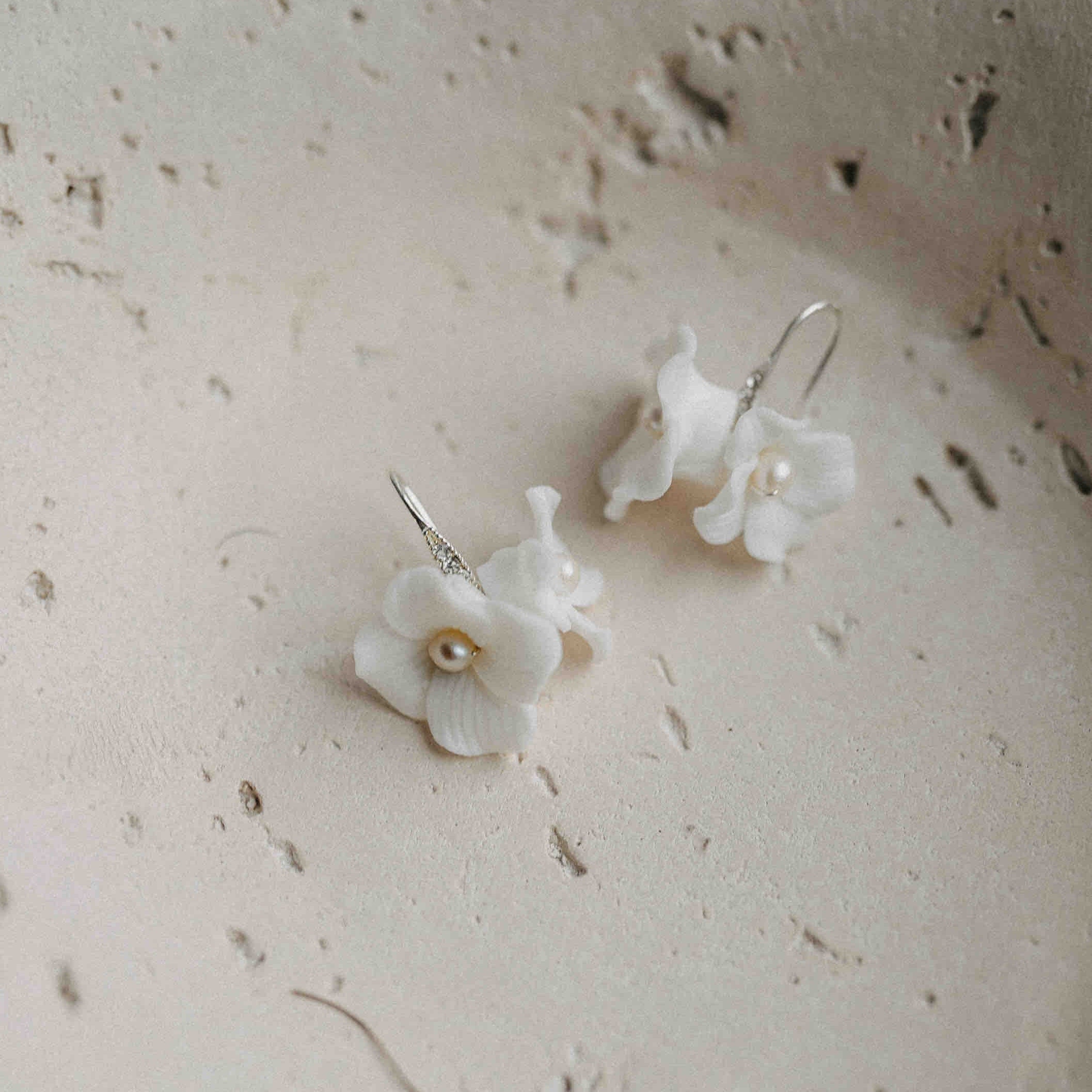 Braut Ohrringe mit Keramik Blüten von Fräulein Maya: Zarte Ohrringe mit kleinen Perlen und Silberhaken, ideal für die Boho-Braut, präsentiert auf strukturierter, cremeweißer Keramik mit natürlichen Sprenkeln und Vertiefungen.