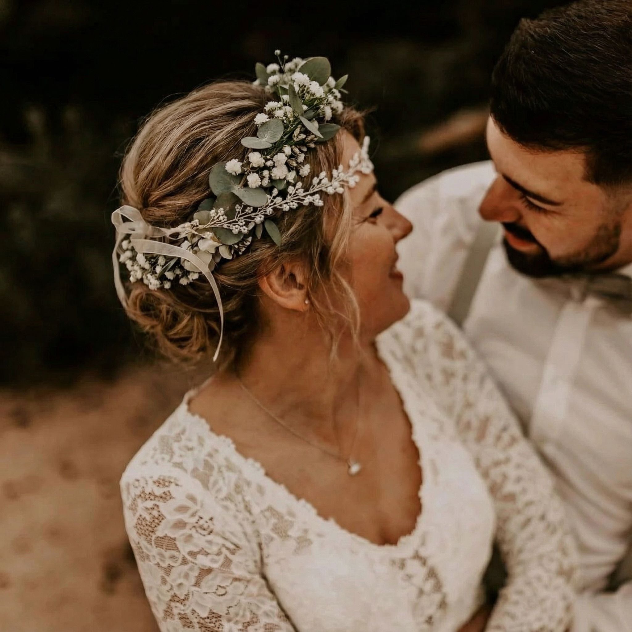 Eine Braut in einem weißen Spitzenkleid und einer Blumenkrone lächelt einen Bräutigam mit Bart und Fliege an. Das Paar schaut sich liebevoll an, umgeben von weichem, warmem Licht und verschwommenem Grün im Hintergrund.