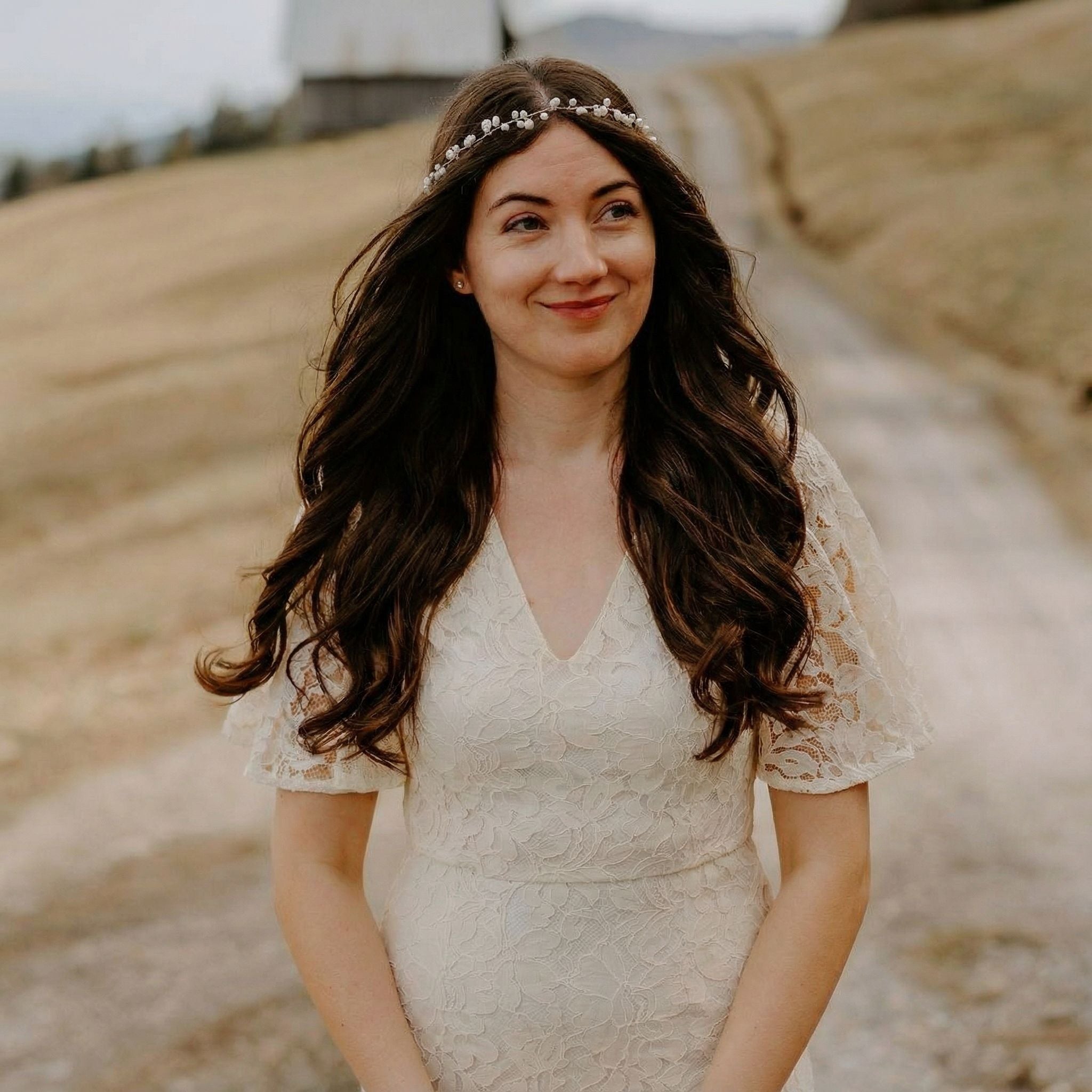 Eine Frau mit langen, gewellten braunen Haaren trägt ein weißes Spitzenkleid mit Flatterärmeln und einem perlenbesetzten Stirnband. Sie steht im Freien auf einem unbefestigten Weg und lächelt leicht. Im Hintergrund sind eine verschwommene ländliche Landschaft und eine Scheune zu sehen.