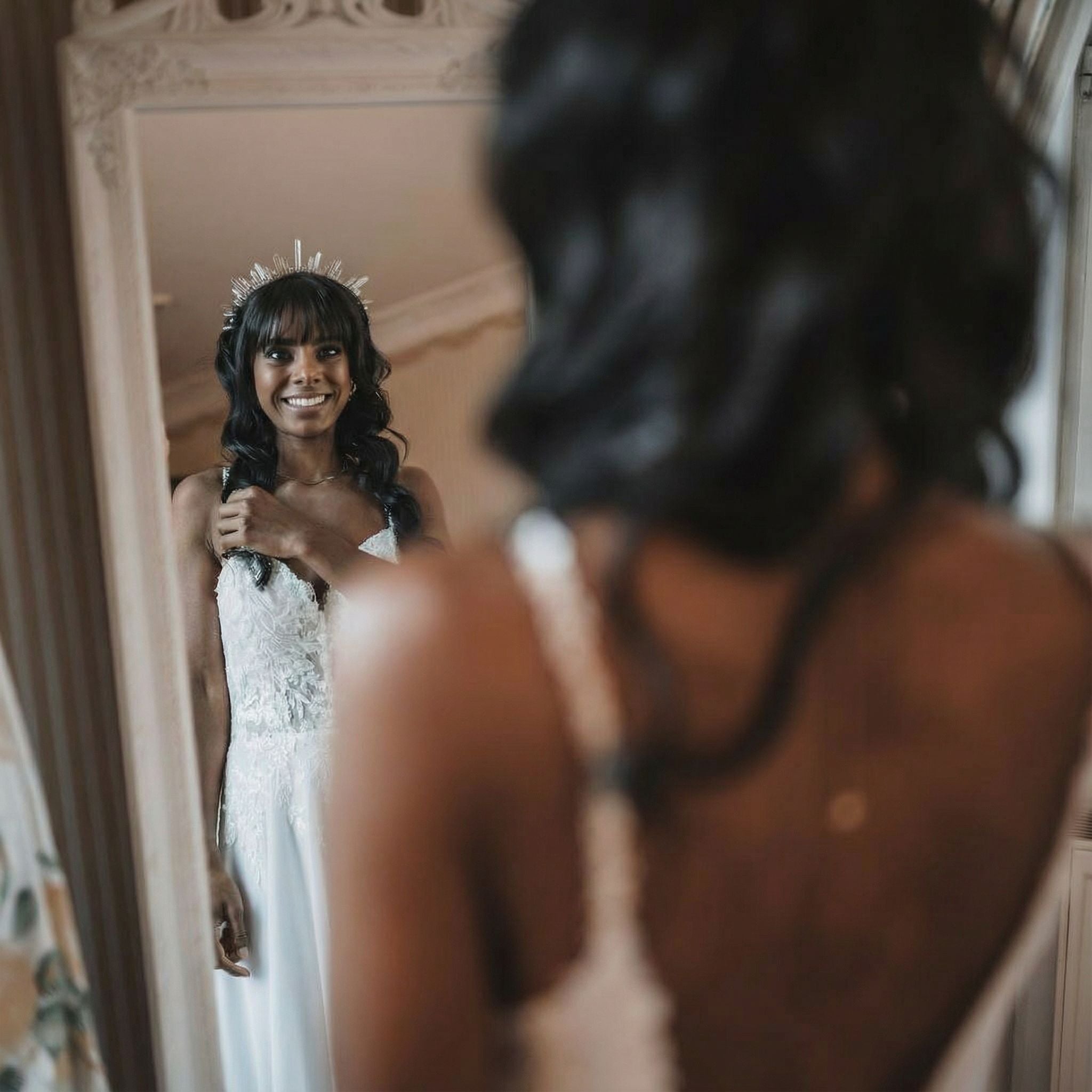 Eine Frau in einem weißen, spitzenbesetzten Hochzeitskleid lächelt, während sie sich in einem Spiegel betrachtet. Sie hat langes dunkles Haar mit Pony und trägt eine kronenartige Kopfbedeckung. Ihre Hand berührt sanft ihr Haar, und die Umgebung ist elegant und sanft beleuchtet.
