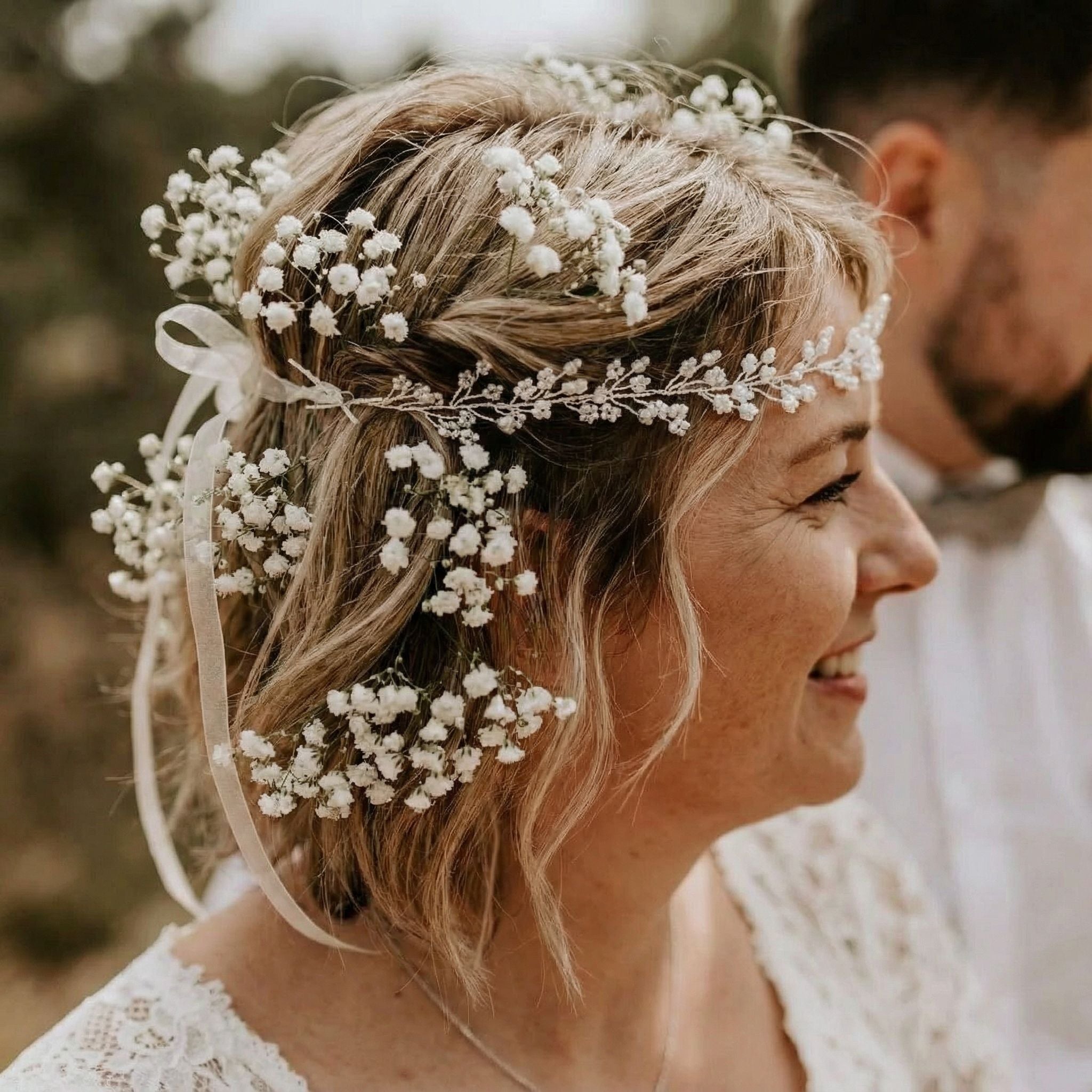 Eine lächelnde Frau mit kurzen blonden Haaren, die mit weißen Schleierkrautblüten und einem weißen Band geschmückt sind, trägt ein weißes Spitzenoberteil. Sie blickt nach rechts, mit einer unscharfen Person und einem Außenhintergrund hinter ihr.