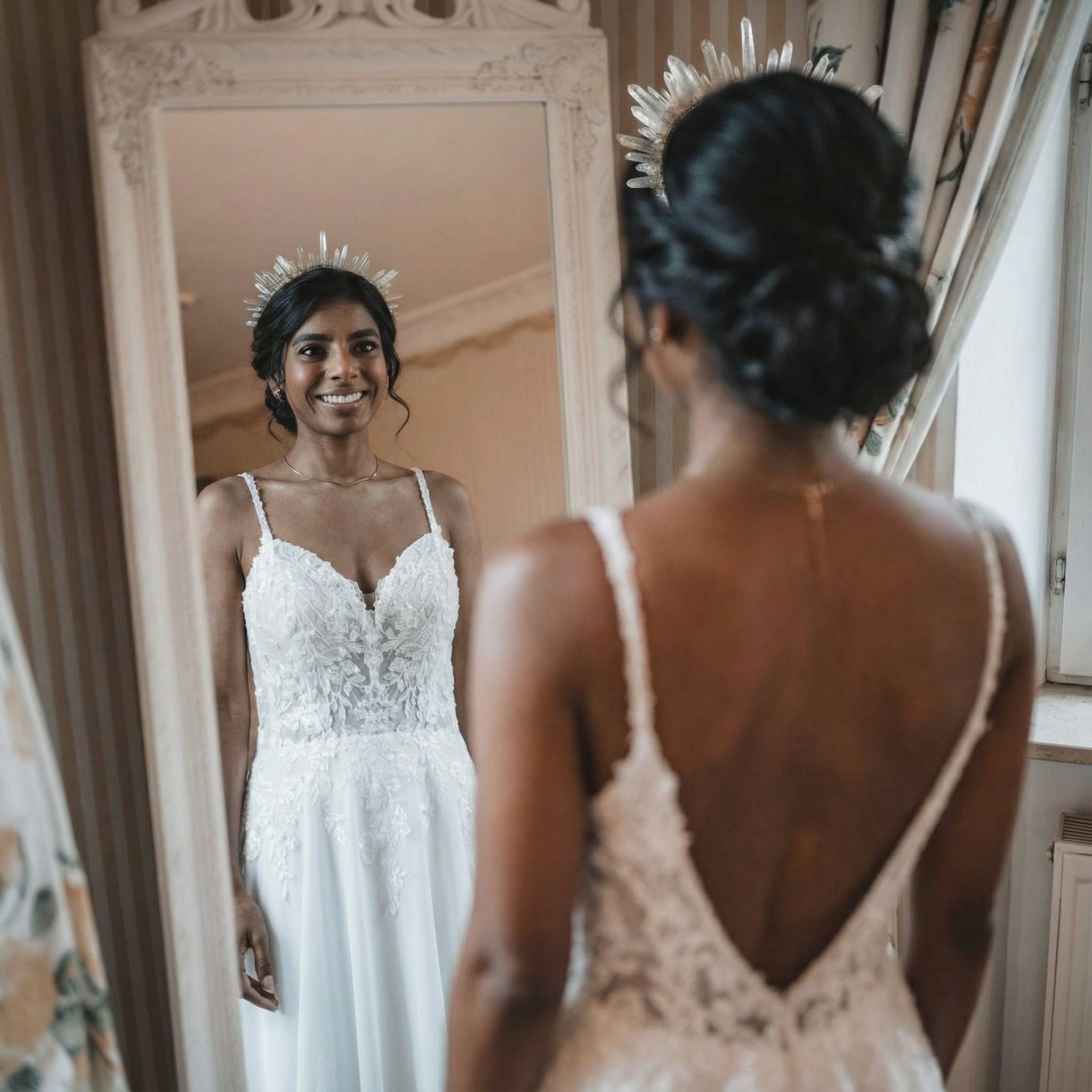 Eine Braut mit dunklem Haar in einer eleganten Hochsteckfrisur und einer Stachelkrone lächelt ihr Spiegelbild in einem Vintage-Spiegel an. Sie trägt ein weißes Spitzenhochzeitskleid mit dünnen Trägern und tiefem Rückenausschnitt und steht in einem klassisch eingerichteten Raum mit gedämpfter Beleuchtung und gemusterten Vorhängen.