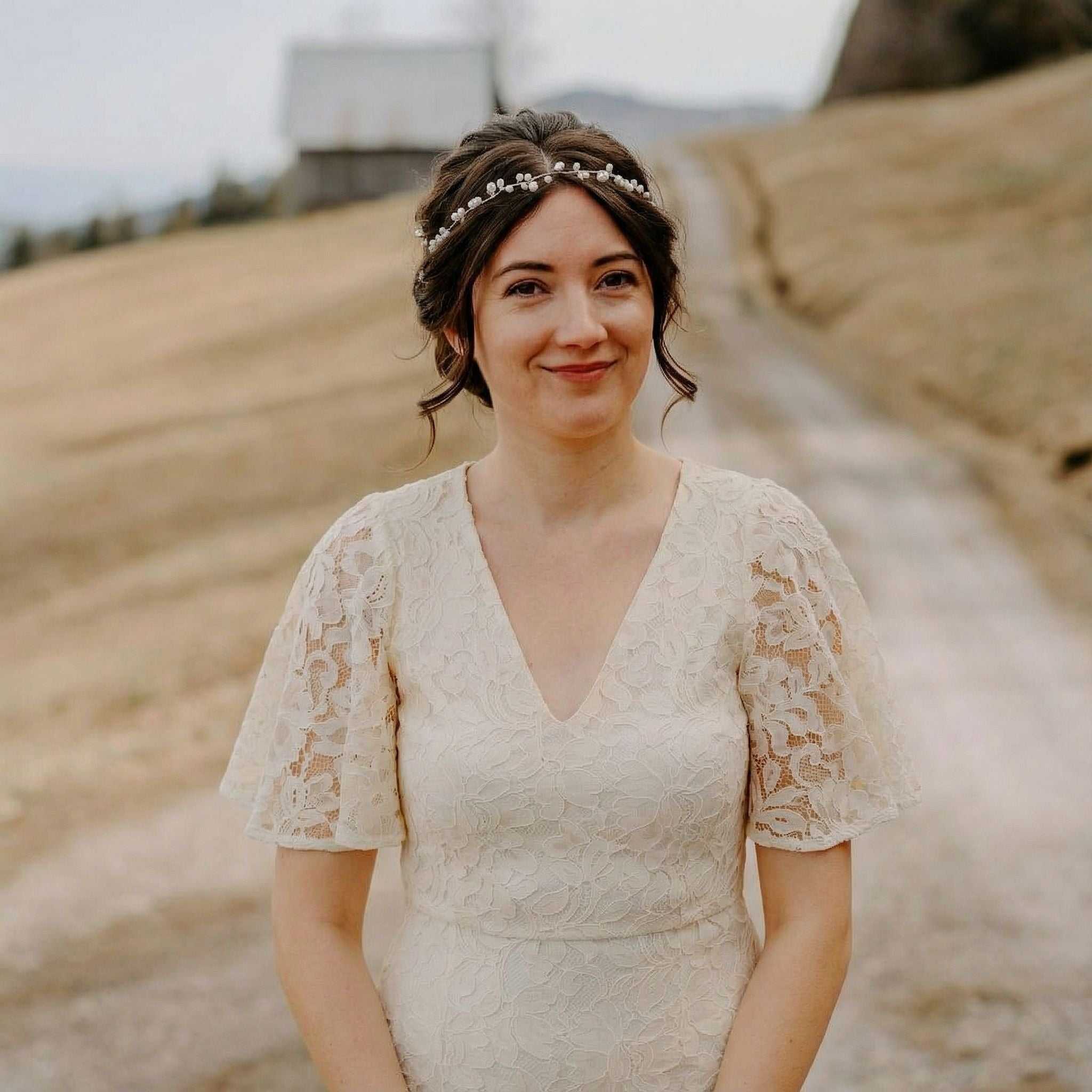 Eine Frau in einem cremefarbenen Spitzenkleid mit Flatterärmeln steht auf einer Landstraße. Sie hat braunes Haar, das mit einem geblümten Stirnband hochgesteckt ist, und lächelt sanft. Im Hintergrund sind eine verschwommene Scheune, grasbewachsene Hügel und ein bedeckter Himmel zu sehen.