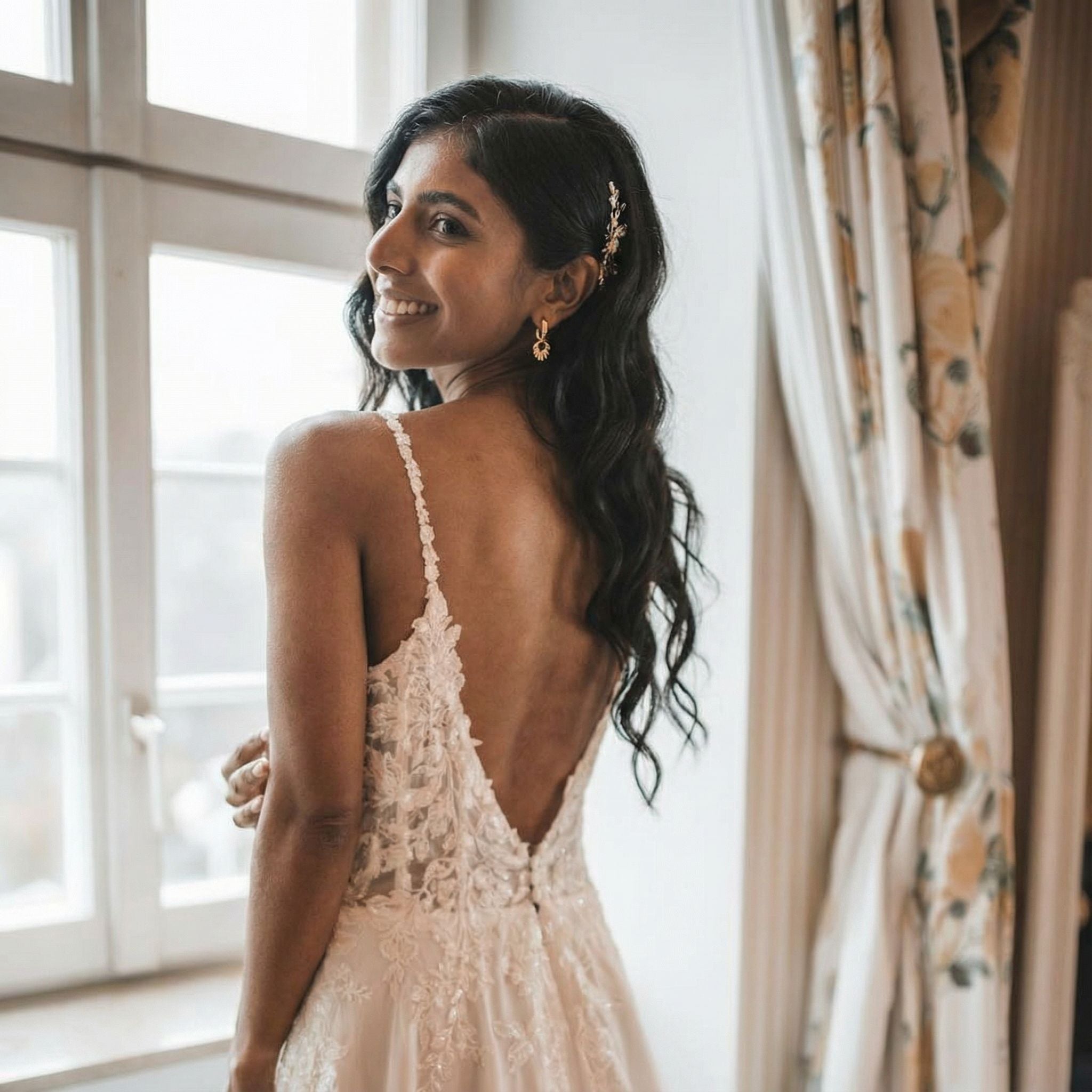 Eine Frau mit langen, gewellten schwarzen Haaren lächelt über ihre Schulter, während sie an einem Fenster steht. Sie trägt ein weißes Spitzenbrautkleid mit dünnen Trägern und tiefem Rückenausschnitt, goldene Ohrringe und einen zarten Haarschmuck. Neben ihr hängen cremefarbene geblümte Vorhänge.