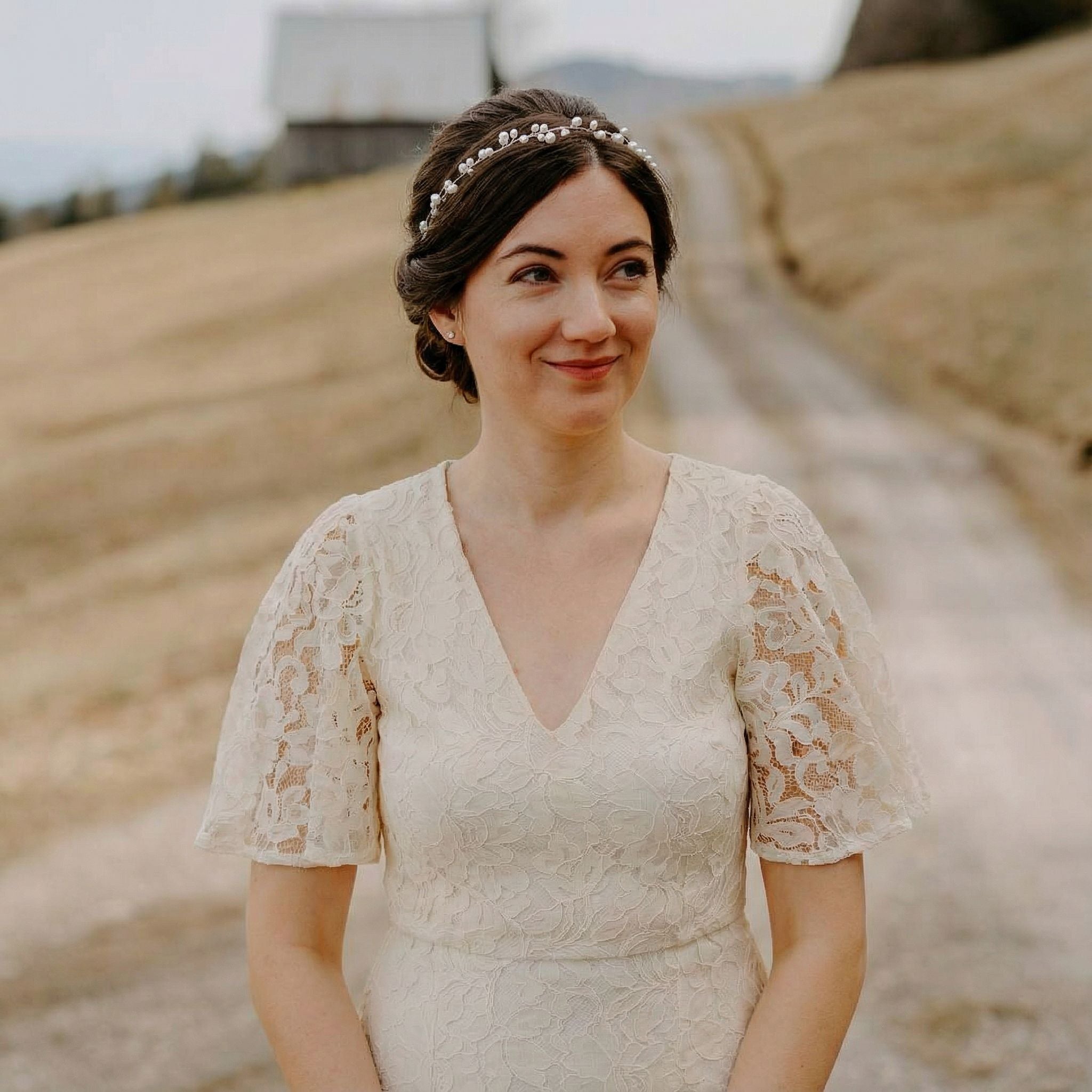 Eine Frau in einem cremefarbenen Spitzenkleid mit Flatterärmeln steht auf einem Feldweg. Sie hat dunkles Haar in einer Hochsteckfrisur mit einem Perlenstirnband und lächelt leicht. Im Hintergrund sind eine verschwommene Scheune und trockene, gebräunte Felder unter einem blassen Himmel zu sehen.