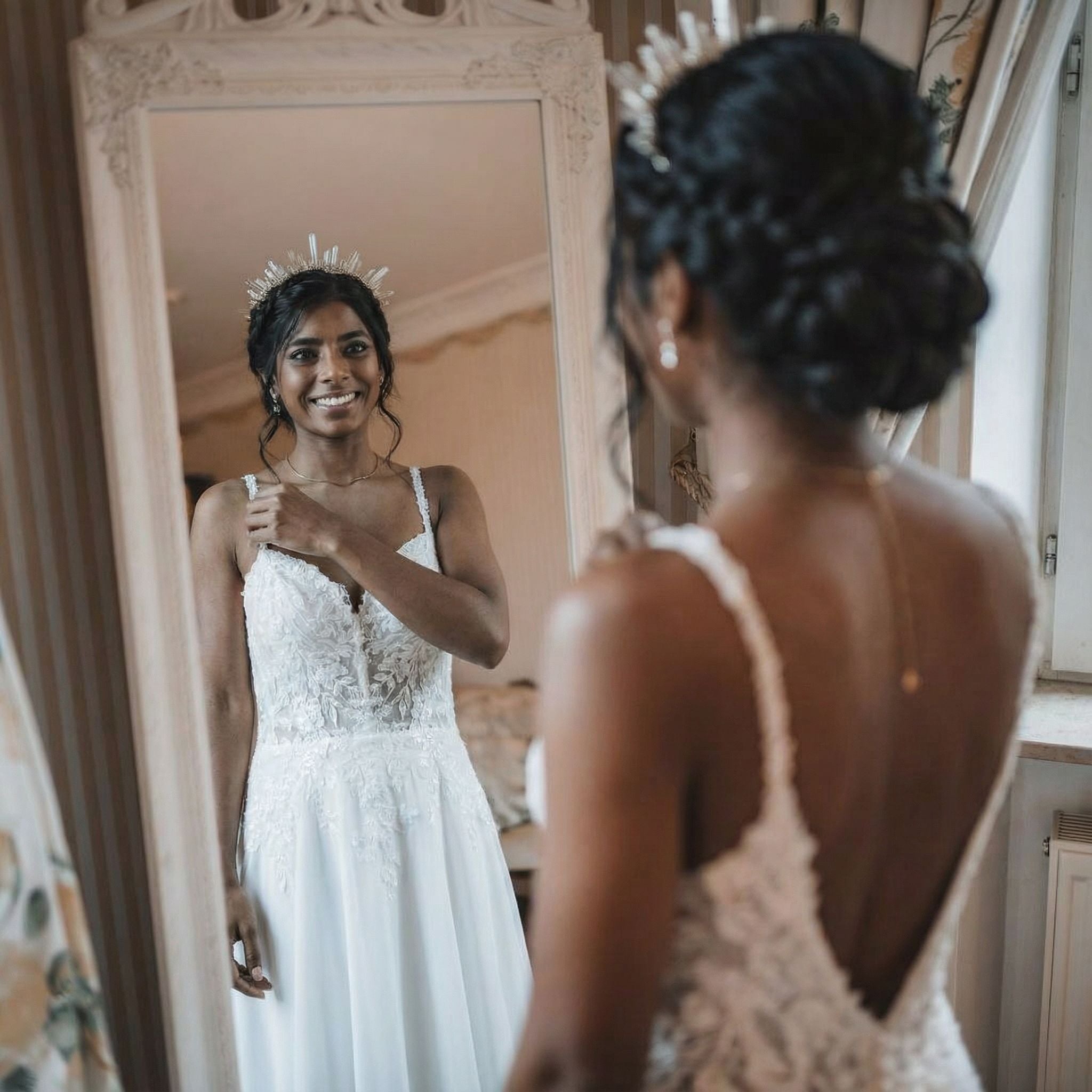 Eine Frau in einem weißen Spitzenhochzeitskleid und einem juwelenbesetzten Kopfschmuck lächelt ihr Spiegelbild an. Ihr dunkles Haar ist zu einer eleganten Hochsteckfrisur gestylt. Der Raum ist mit sanfter Beleuchtung, geblümten Vorhängen und kunstvollem Dekor ausgestattet, was eine warme, feierliche Atmosphäre schafft.