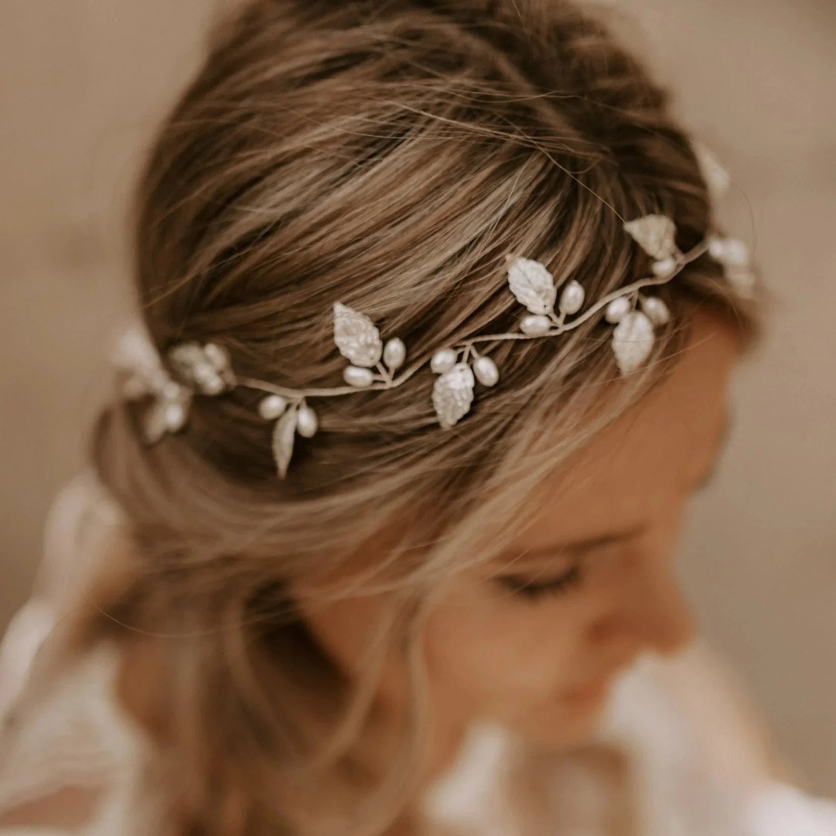 Eine Braut in einem langärmeligen Spitzenkleid und Kopfschmuck wird von einem Bräutigam in einem weißen Hemd umarmt und am Hals geküsst, während sie auf einer Sandfläche mit Bäumen im Hintergrund steht.