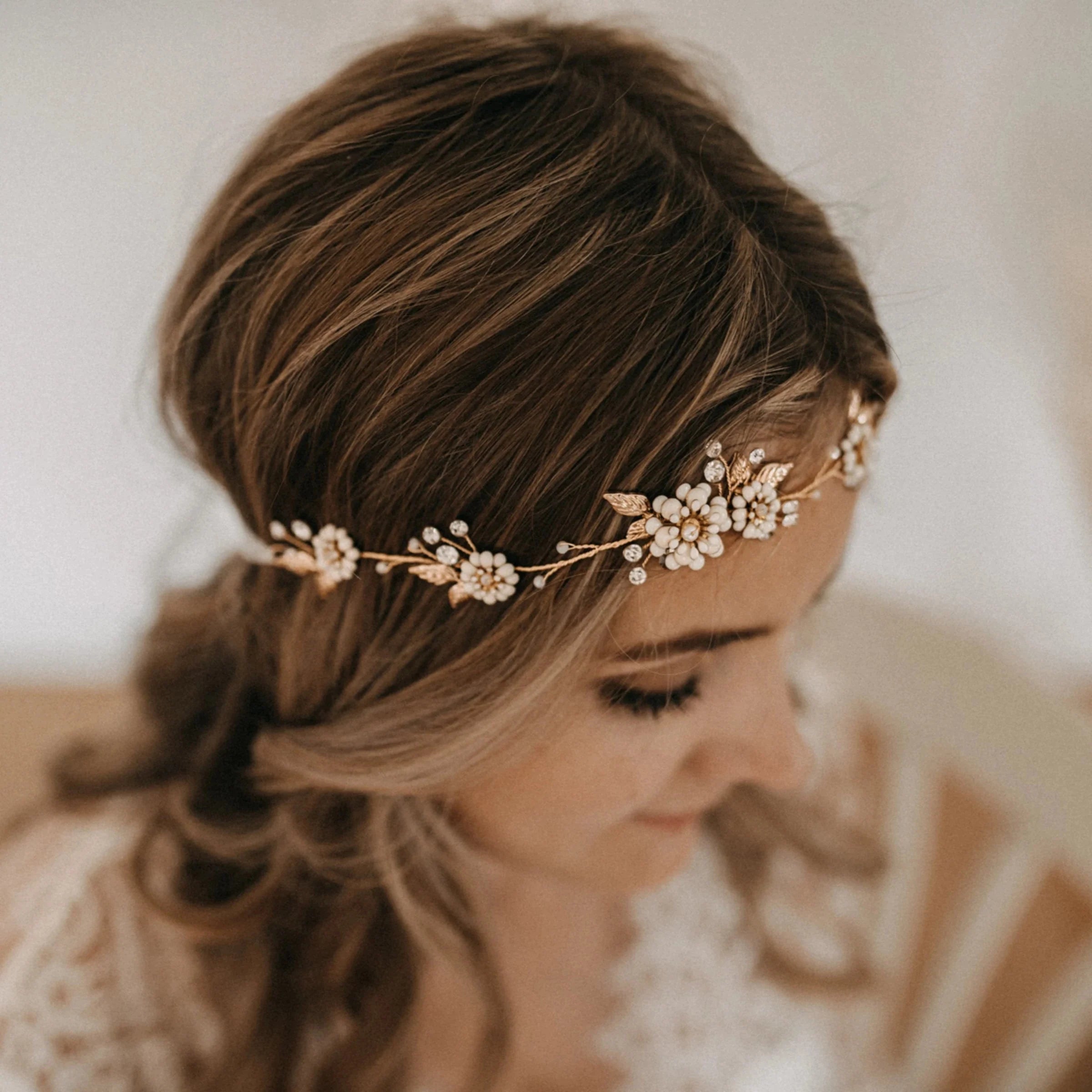 Eine Frau mit gewelltem braunem Haar trägt einen zarten goldenen Blumenschmuck mit Perlen und Kristallen. Sie blickt nach unten, und ihre komplizierte Frisur ist teilweise sichtbar. Der Hintergrund ist sanft verschwommen.