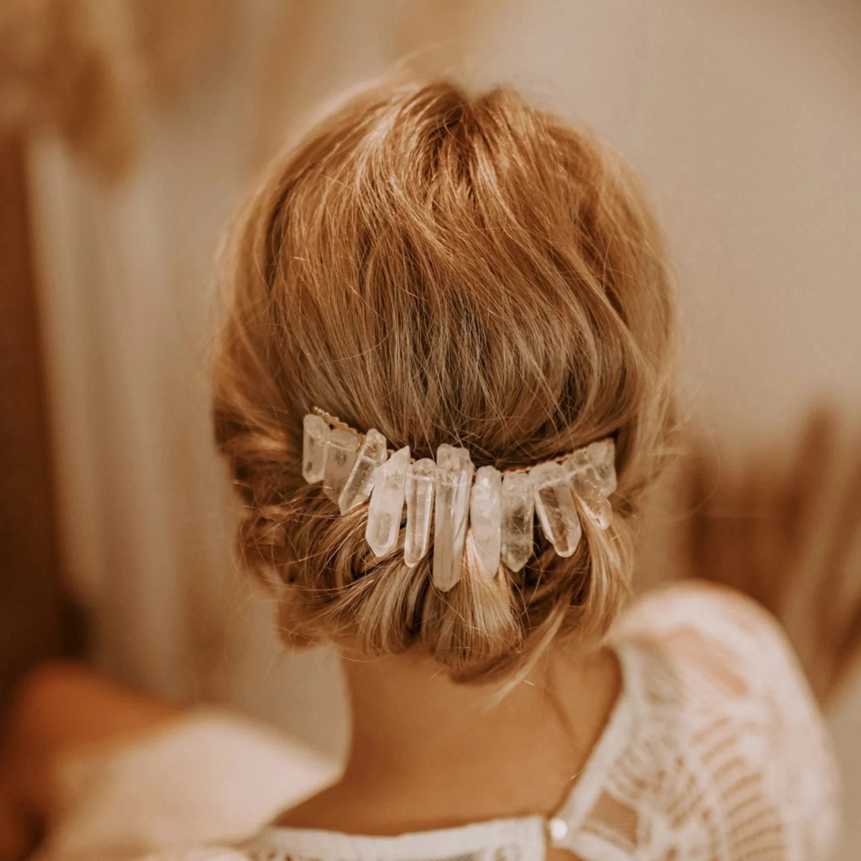 Eine Frau mit blondem, zu einer Hochsteckfrisur frisiertem Haar, das mit einem dekorativen Haarkamm mit klaren Kristallspitzen geschmückt ist. Sie trägt ein weißes Spitzenkleid mit verschlungenen Mustern.