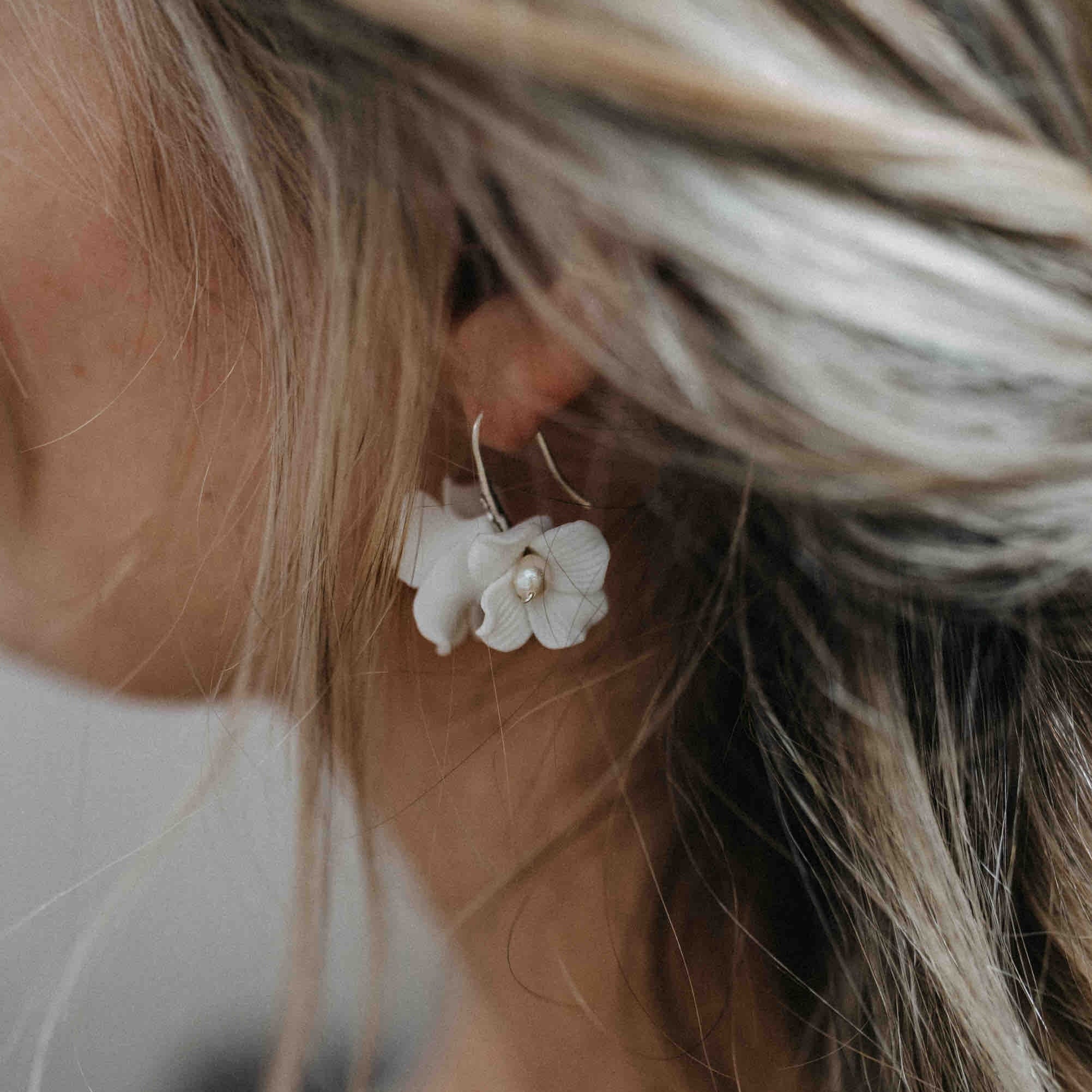 Nahaufnahme eines Frauenohrs mit einem großen weißen blumenförmigen Ohrring mit einer Perle in der Mitte. Ihr blondes Haar ist locker gestylt, mit weichen, natürlichen Strähnchen und Strähnen, die ihr Gesicht umrahmen.