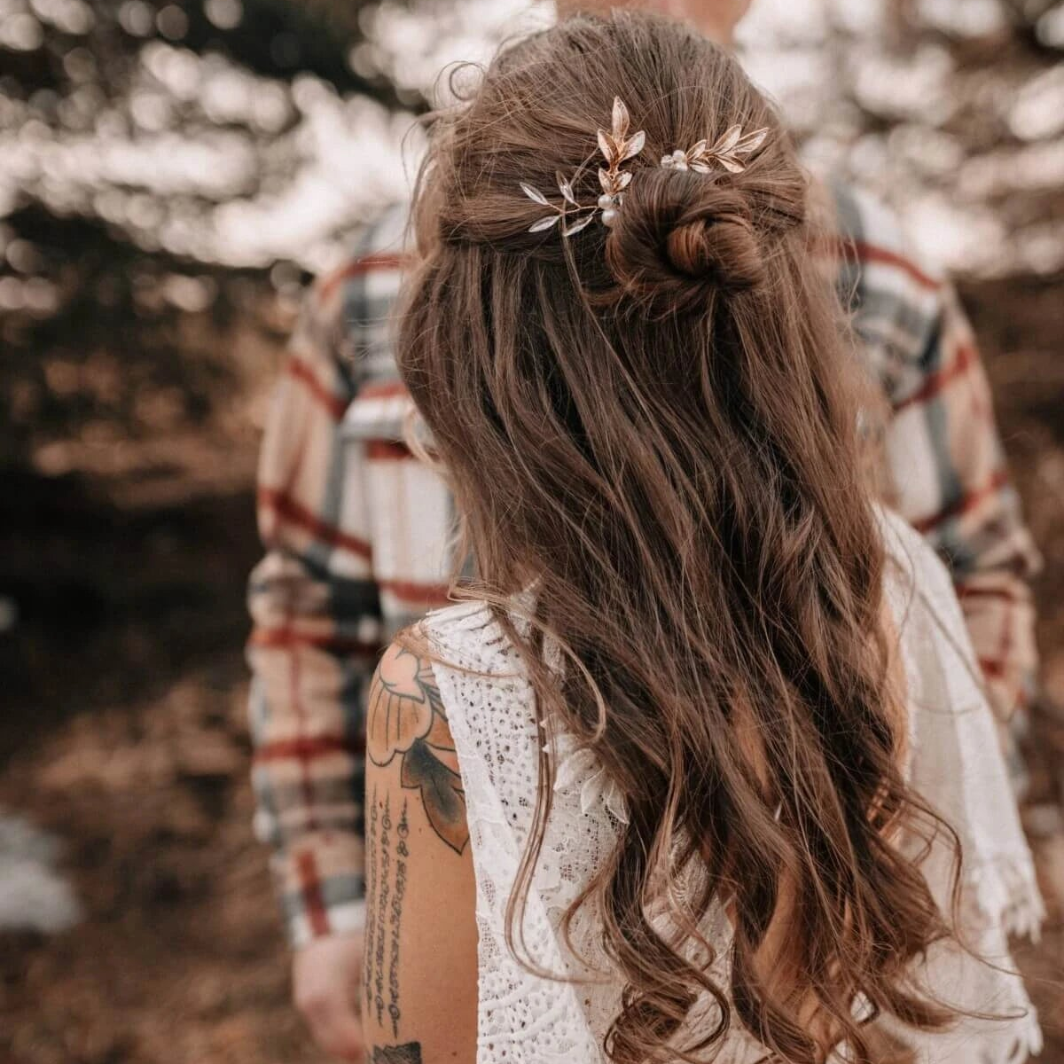 Eine Frau mit langen, gewellten braunen Haaren, die zu einem halb hochgesteckten Dutt mit einer dekorativen Haarnadel frisiert sind, steht im Freien. Sie trägt ein weißes Spitzentop und hat ein Blumentattoo auf dem Oberarm. Im unscharfen Hintergrund steht eine Person in einem karierten Hemd.