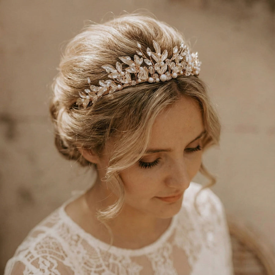 Eine Frau mit blondem Haar, das zu einer lockeren Hochsteckfrisur frisiert ist, trägt einen zarten Kopfschmuck mit Perlen und Kristallblättern. Sie hat weiche Locken, die ihr Gesicht umrahmen, und trägt ein weißes Spitzenkleid. Sie blickt mit heiterem Blick auf einen neutralen Hintergrund.