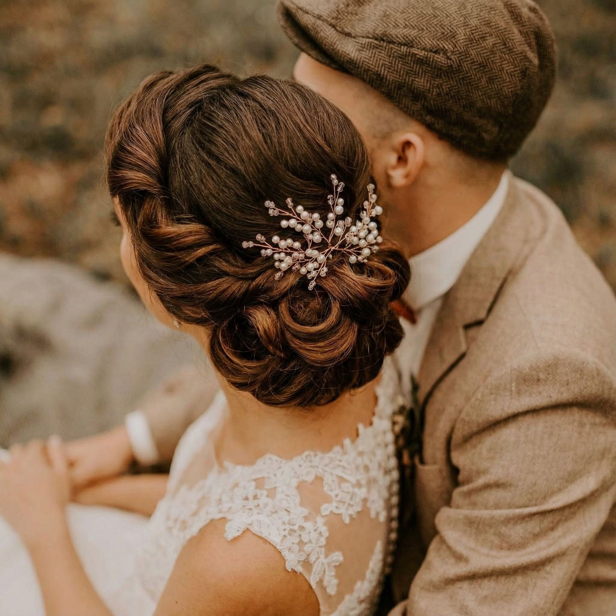 Eine Frau in einem weißen Spitzenkleid mit einer eleganten geflochtenen Hochsteckfrisur, die mit Perlenhaarnadeln verziert ist, sitzt dicht neben einem Mann in einem braunen Tweedanzug und einer flachen Kappe. Beide befinden sich im Freien, und ihre Gesichter sind von der Kamera abgewandt, was eine intime Atmosphäre schafft.