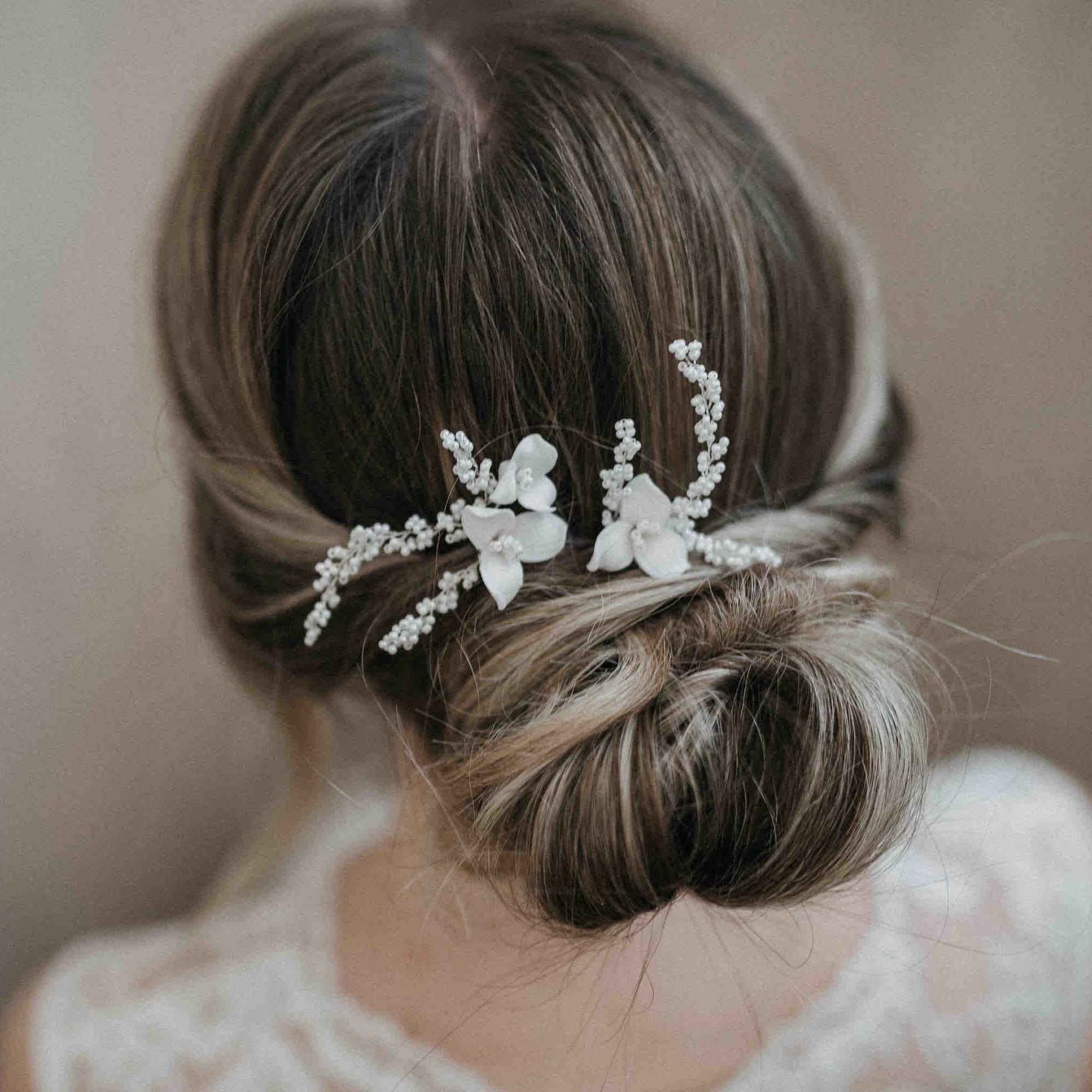 Eine Frau mit braunem Haar in einem lockeren, eleganten Dutt, geschmückt mit Fräulein Maya Keramik Haarnadeln mit Blumen und feinen Perlen (2 Stück). Die Rückseite ihres Spitzenkleides ist vor einem sanft verschwommenen Hintergrund teilweise sichtbar.