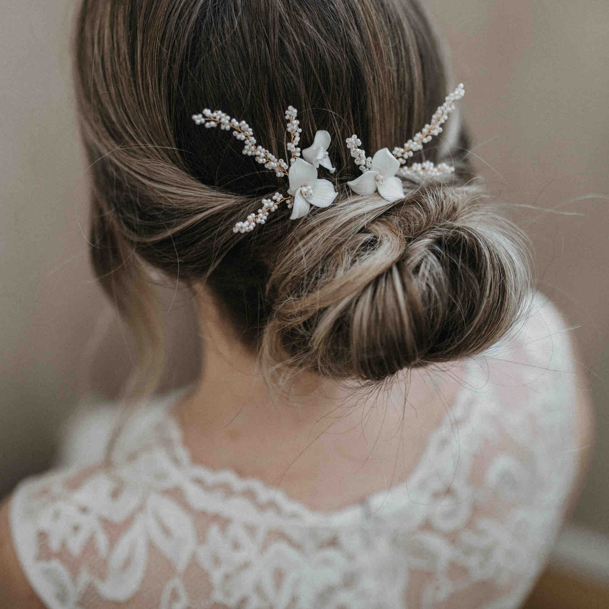 Eine Frau mit braunem Haar, das zu einem tiefen Dutt gestylt ist, trägt die Keramik Haarnadeln mit Blumen und feinen Perlen (2 Stück) von Fräulein Maya. Zarte weiße Keramikblumen und Perlen betonen ihre elegante Frisur und ergänzen perfekt ihr Spitzenkleid.