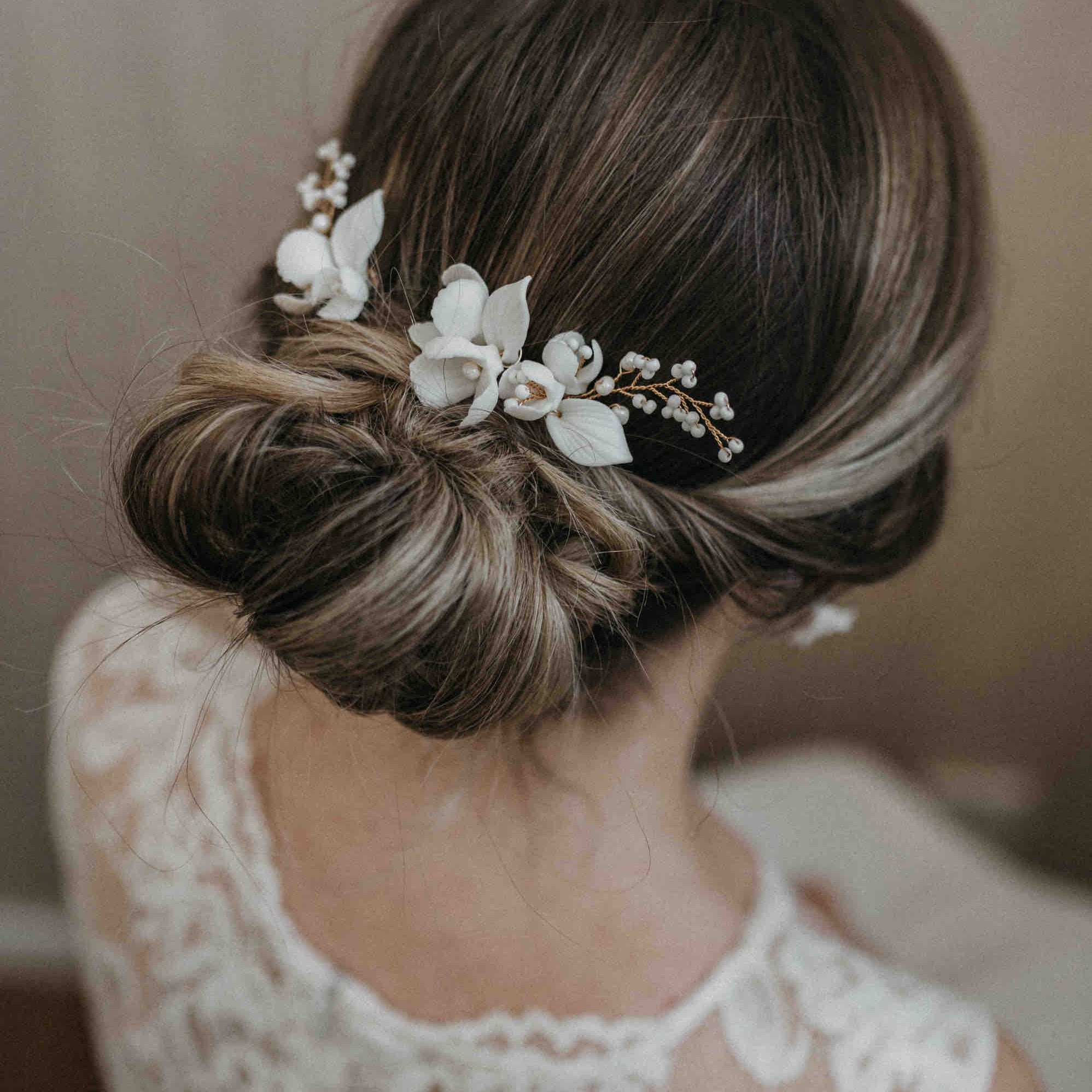 Eine Frau mit braunem Haar in einem eleganten tiefen Dutt, geschmückt mit Fräulein Maya Haarnadeln mit großen Keramik Blumen (3 Stück) und Perlenakzenten, trägt ein Spitzenkleid. Das Foto von hinten hebt ihre aufwendige Brautfrisur und ihre Accessoires hervor.
