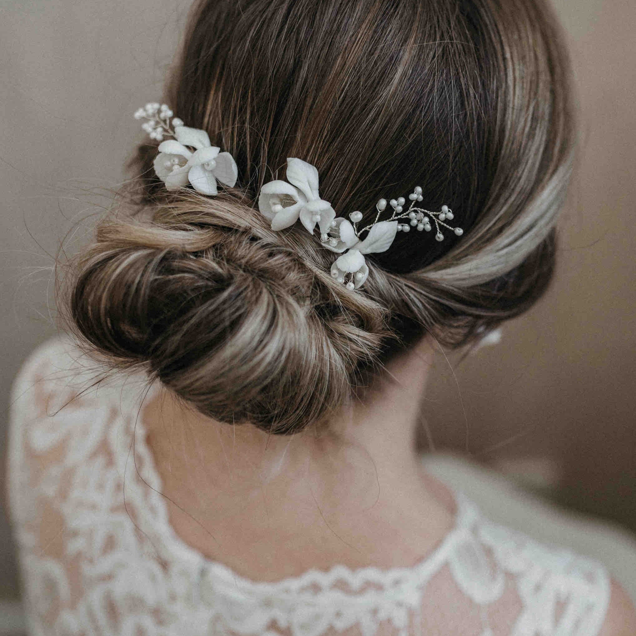 Eine Frau mit braunem Haar, das zu einem niedrigen geflochtenen Dutt gestylt ist, geschmückt mit Fräulein Maya Haarnadeln mit großen Keramik Blumen (3 Stück) und weißen Blumen. Sie trägt ein Spitzenkleid, das von hinten fotografiert wurde, um ihre elegante Brautfrisur zur Geltung zu bringen.