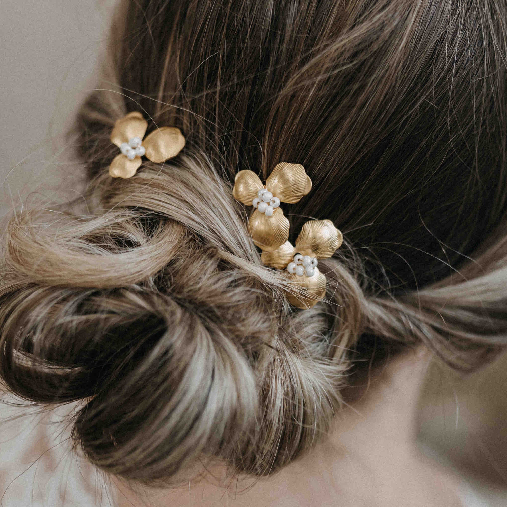 Eine Nahaufnahme des hellbraunen Haars einer Frau in einem lockeren, niedrigen Dutt, gestylt mit Fräulein Maya Haarnadeln mit goldenen Blüten (3 Stück) - elegante goldene Blumenhaarnadeln mit weißen Perlen in der Mitte. Der Hintergrund ist sanft verschwommen.